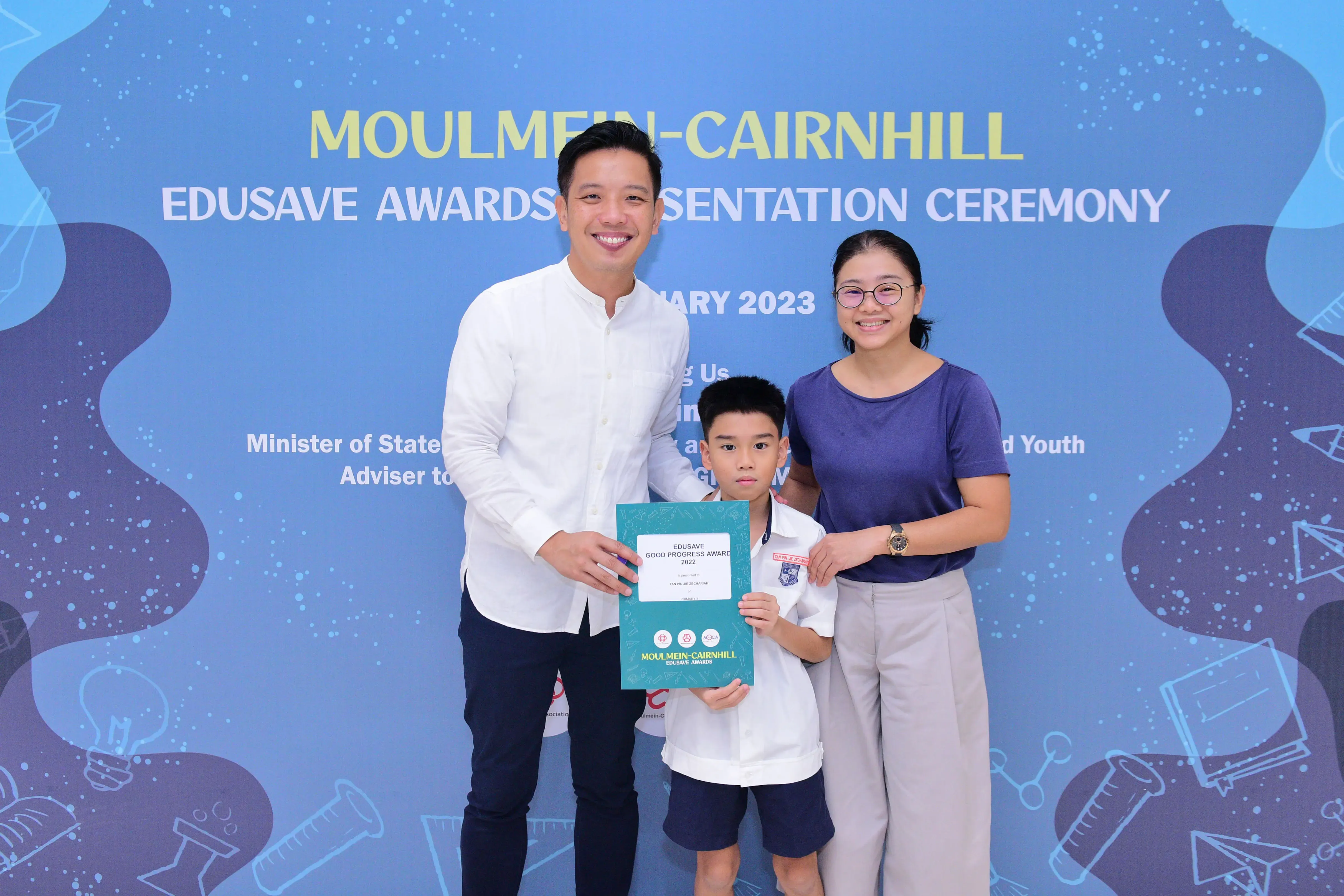 A boy in school uniform holds an Edusave award certificate, flanked by two smiling adults at a 2023 ceremony.