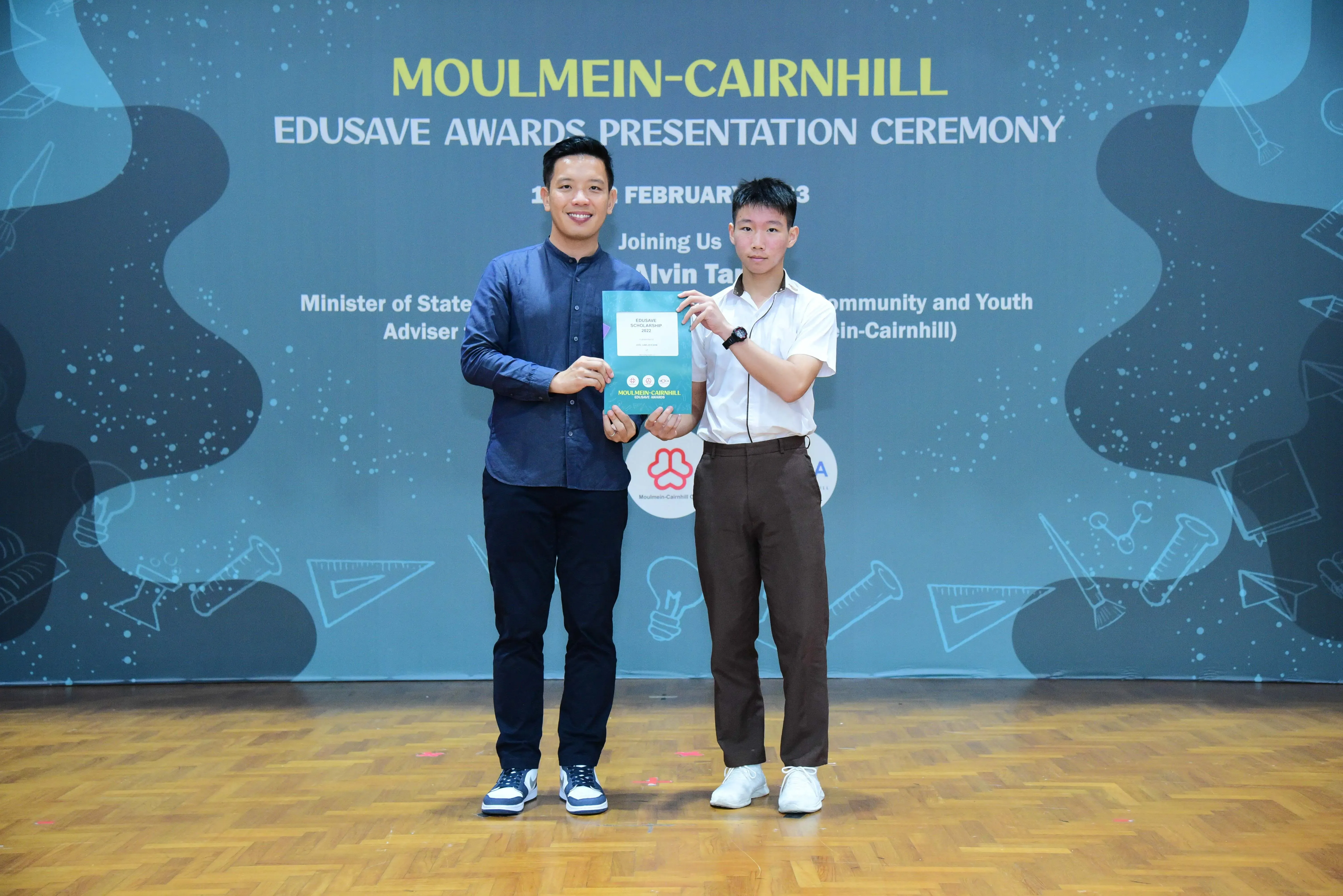 Two men hold a certificate together at the Moulmein-Cairnhill Edusave Awards Presentation Ceremony backdrop.
