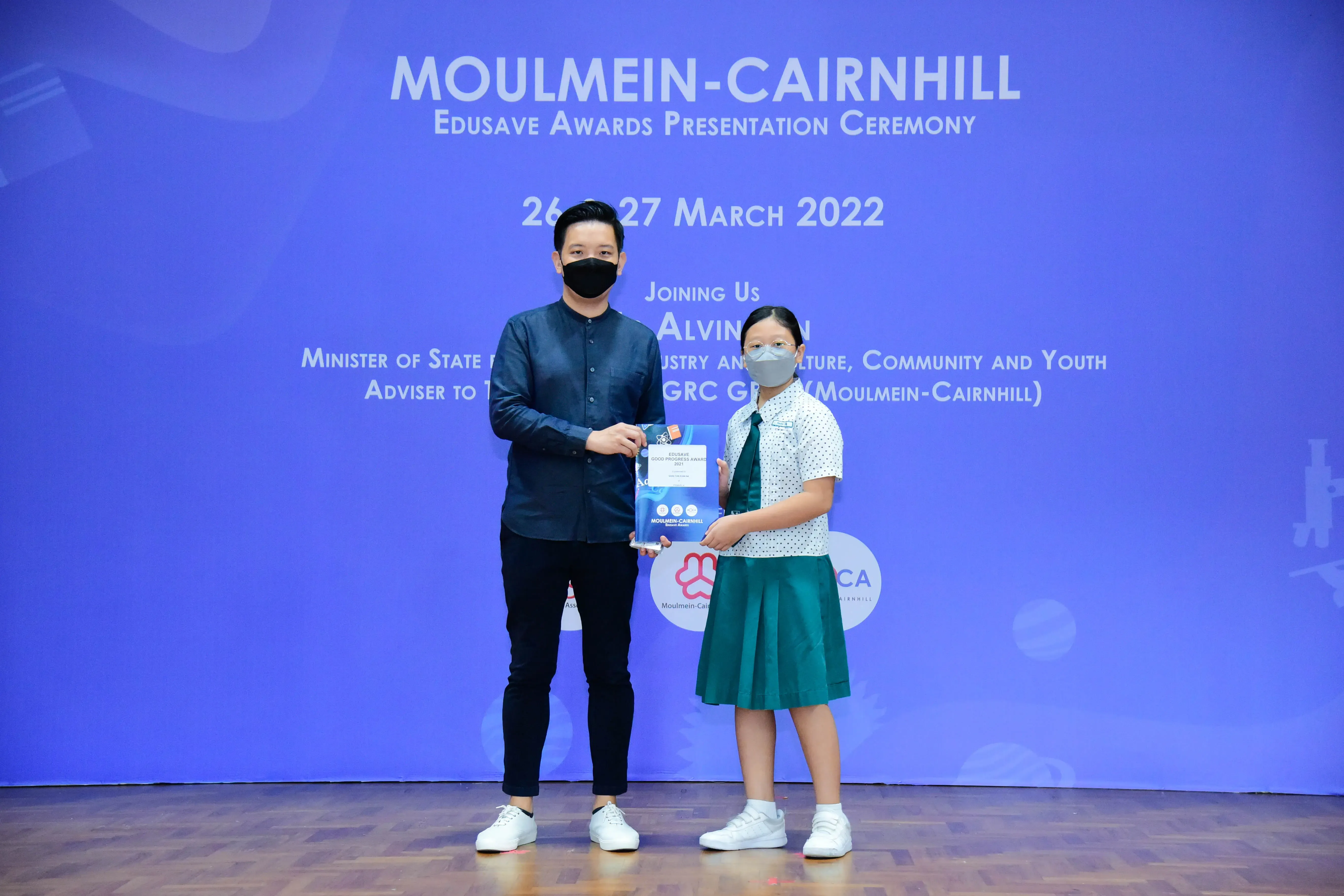 A masked adult and a young student in uniform hold an award at the Moulmein-Cairnhill Edusave Awards Ceremony 2022.