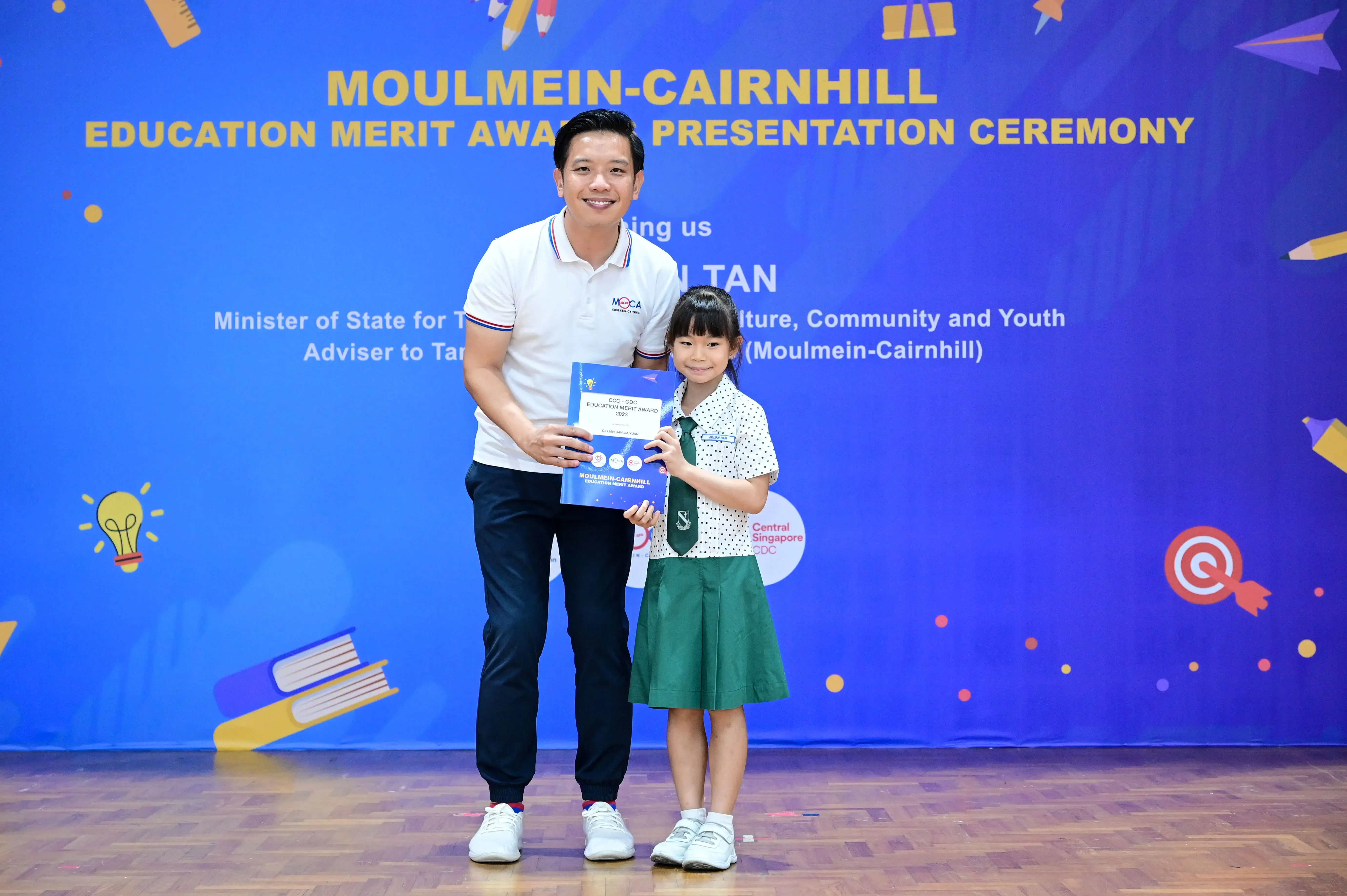 A man and a young girl in school uniform hold an award booklet at the Moulmein-Cairnhill Education Merit Award Ceremony.