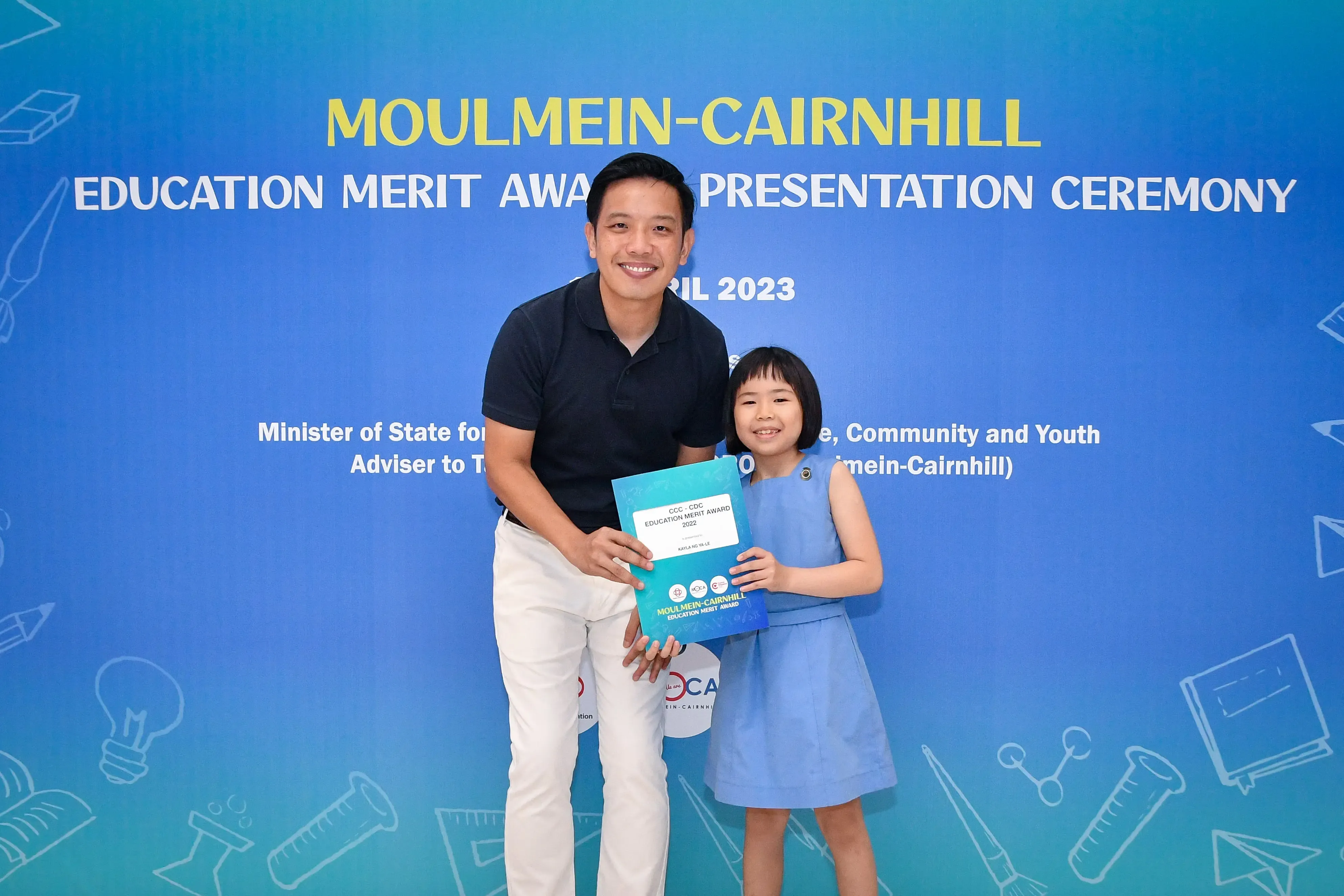 A man and a young girl hold an award certificate at the 2023 Moulmein-Cairnhill Education Merit Award Ceremony.