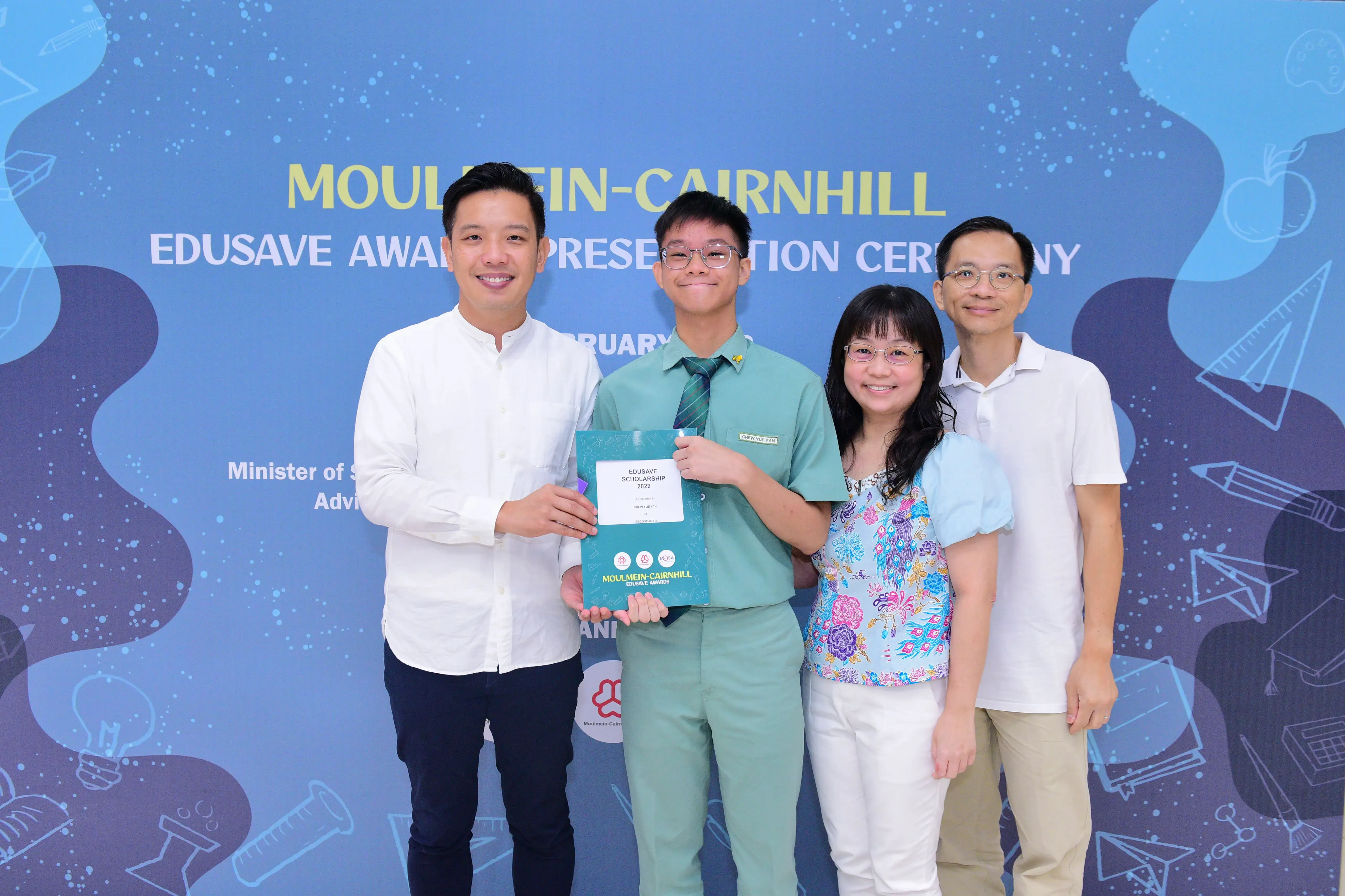 Four people smile at the Moulmein-Cairnhill Edusave Award Presentation Ceremony, with a student holding a scholarship folder.