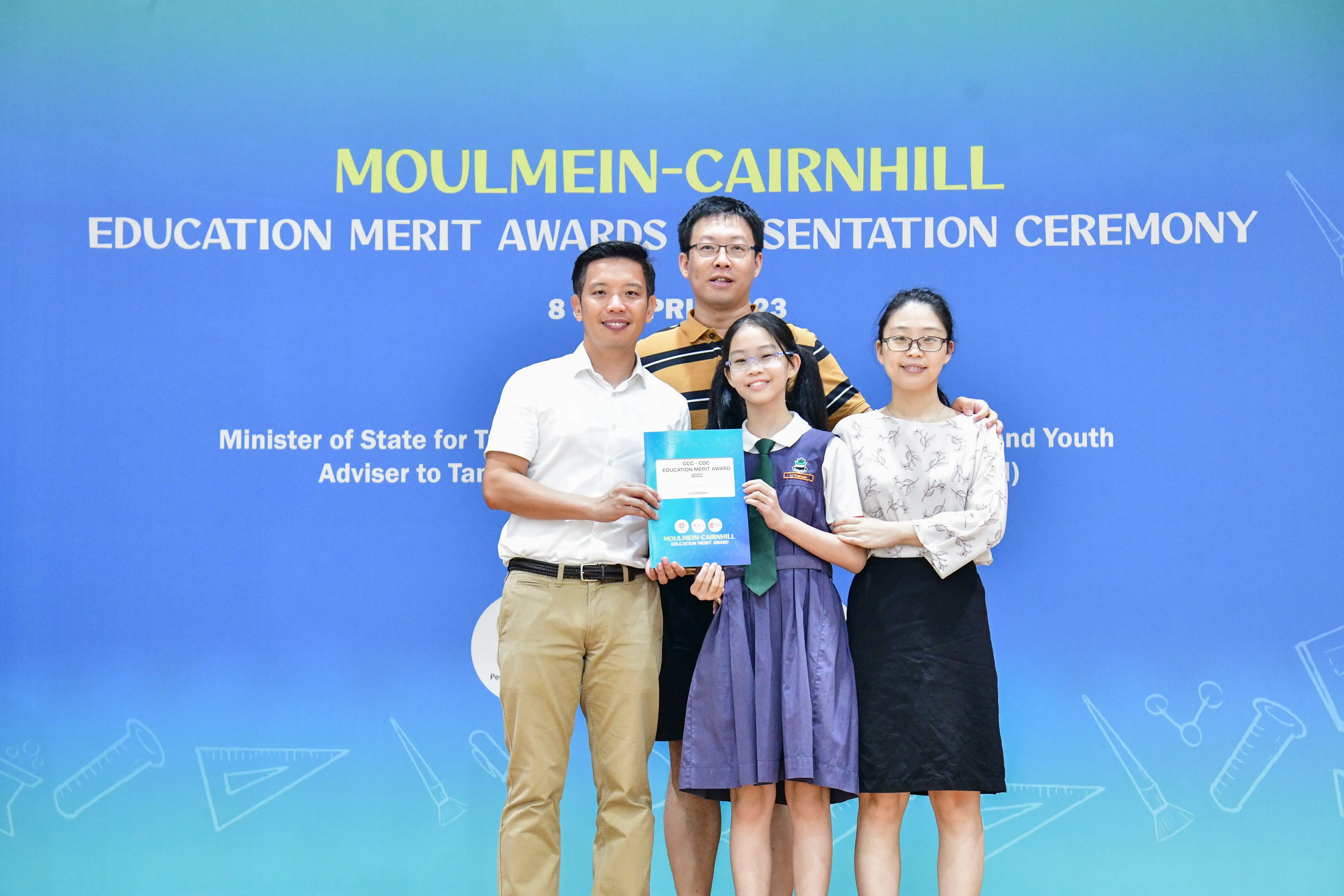 Four people pose before a Moulmein-Cairnhill Education Merit Awards banner, with a young girl holding a blue award certificate.