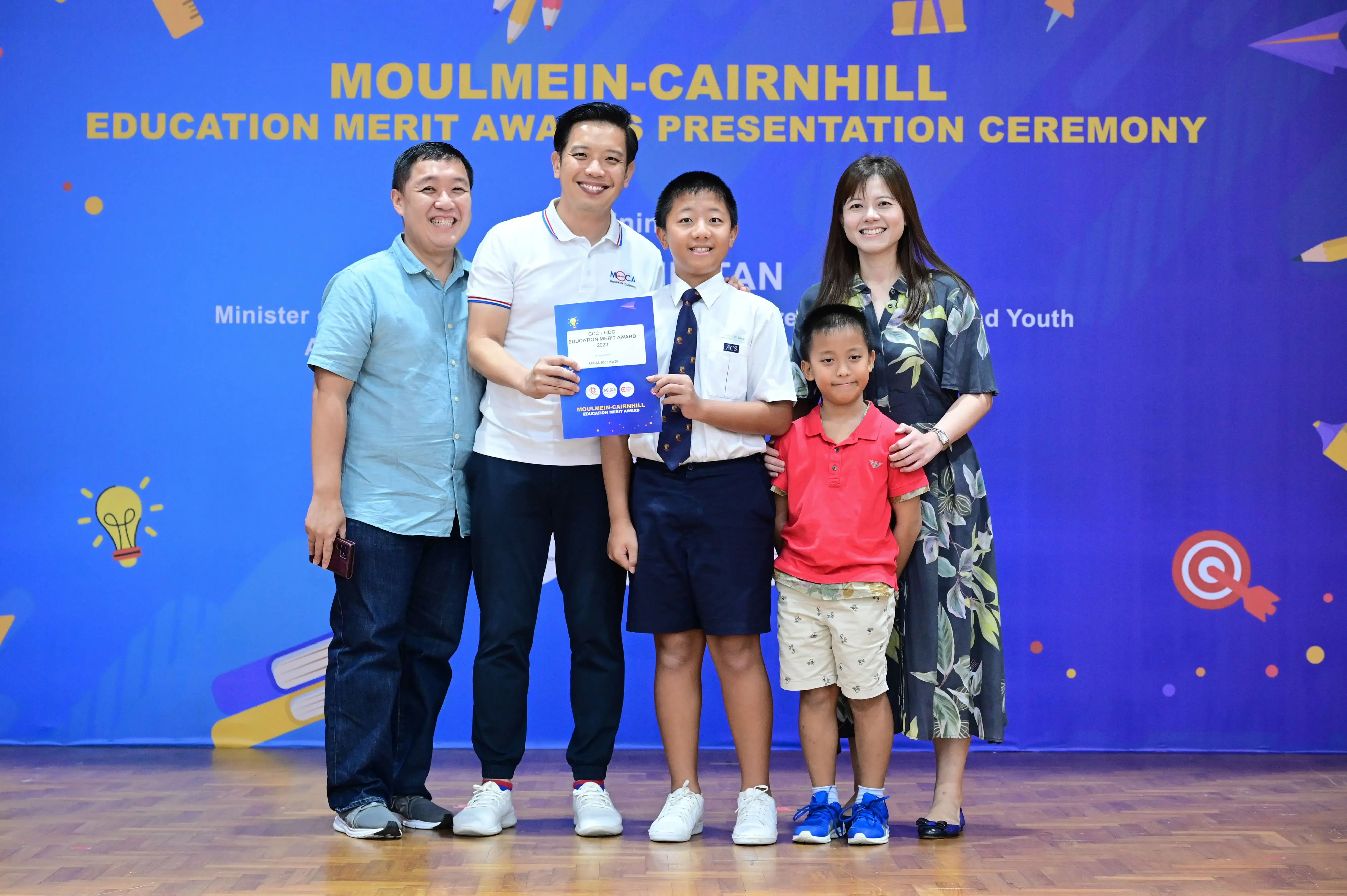 Five people pose at the Moulmein-Cairnhill Education Merit Awards Presentation Ceremony, with a boy holding a blue award certificate.