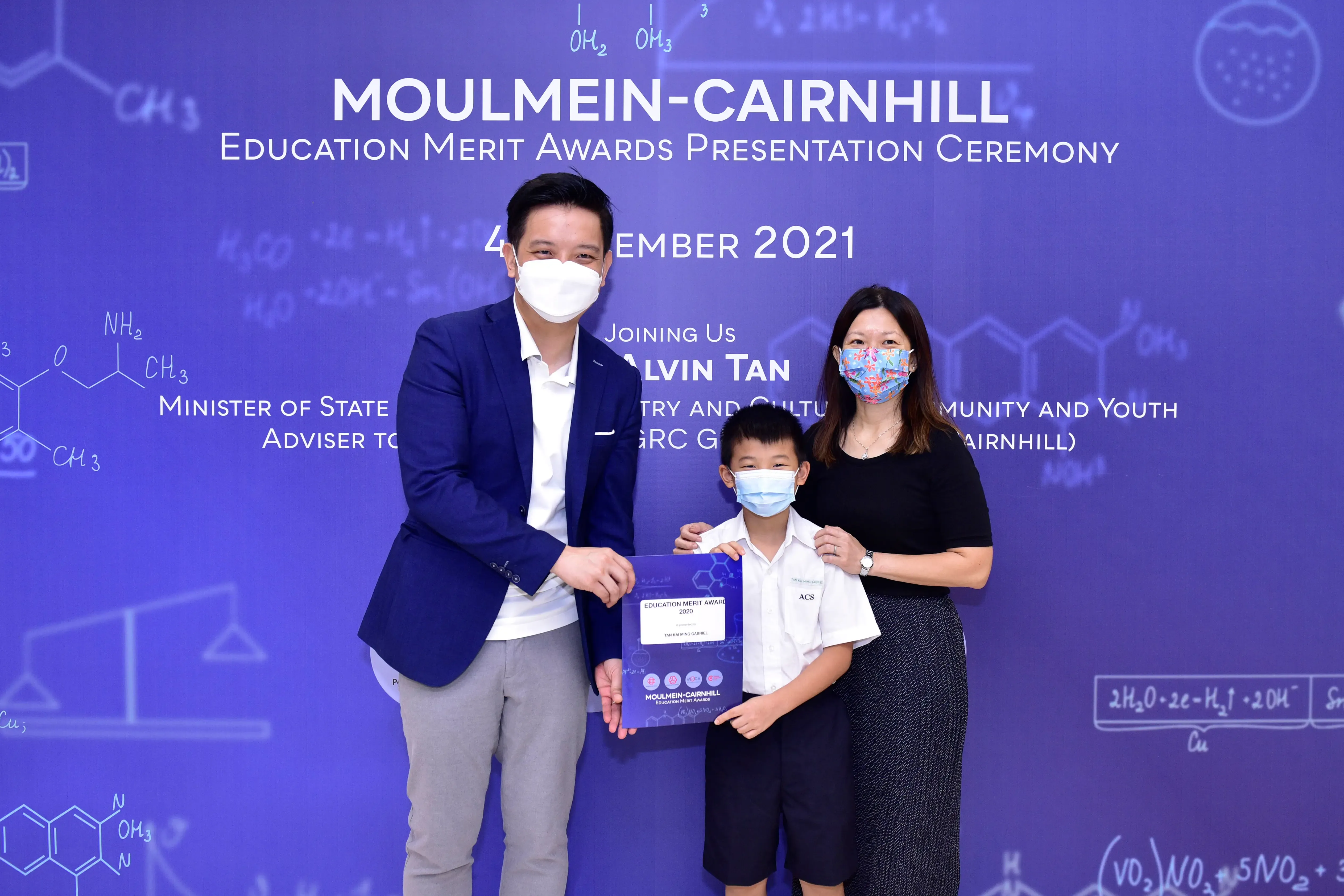 Two masked adults and a masked boy hold an award certificate at the Moulmein-Cairnhill Education Merit Awards 2021.