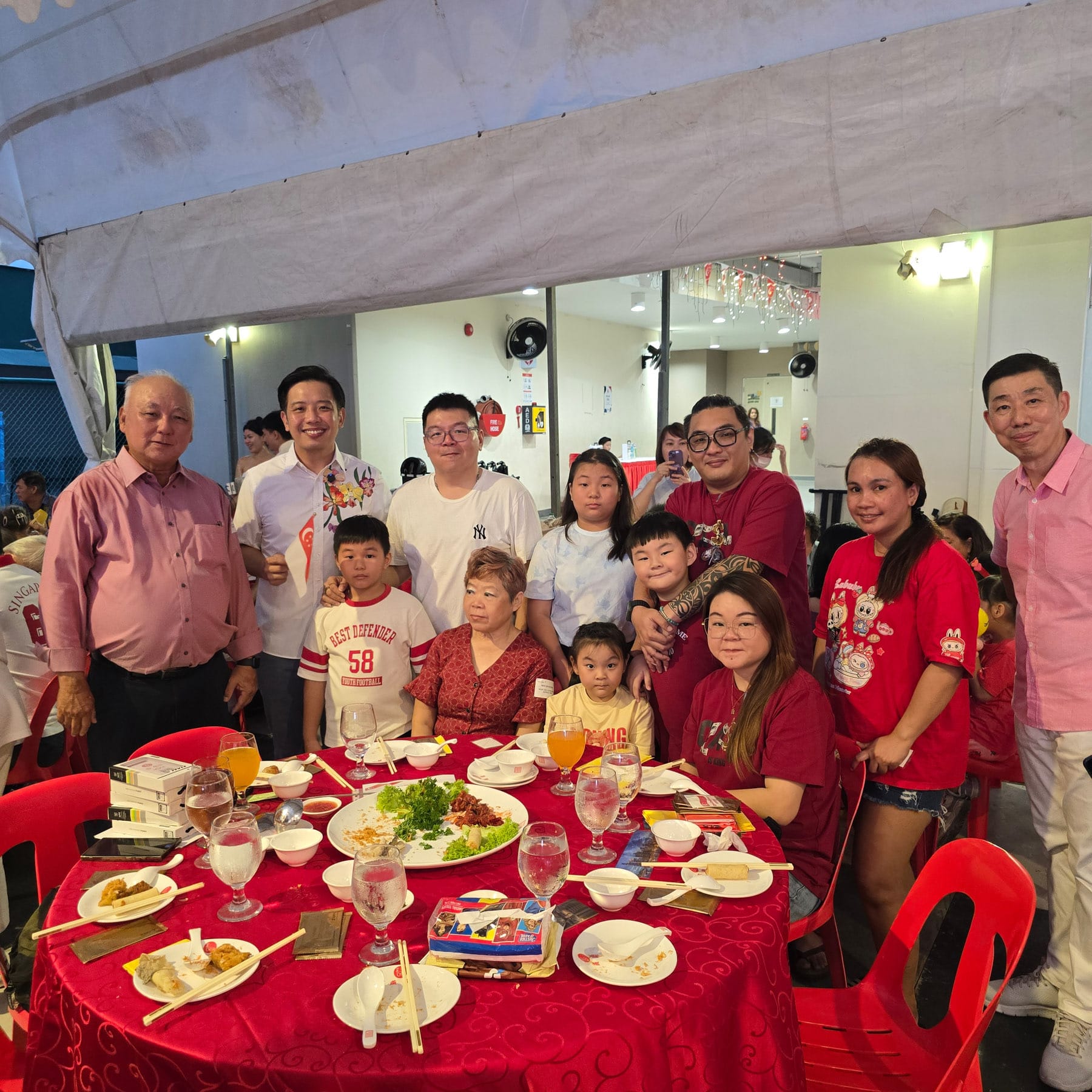 A group of adults and children pose together at a round red table with food, chopsticks, and drinks at a festive dinner.