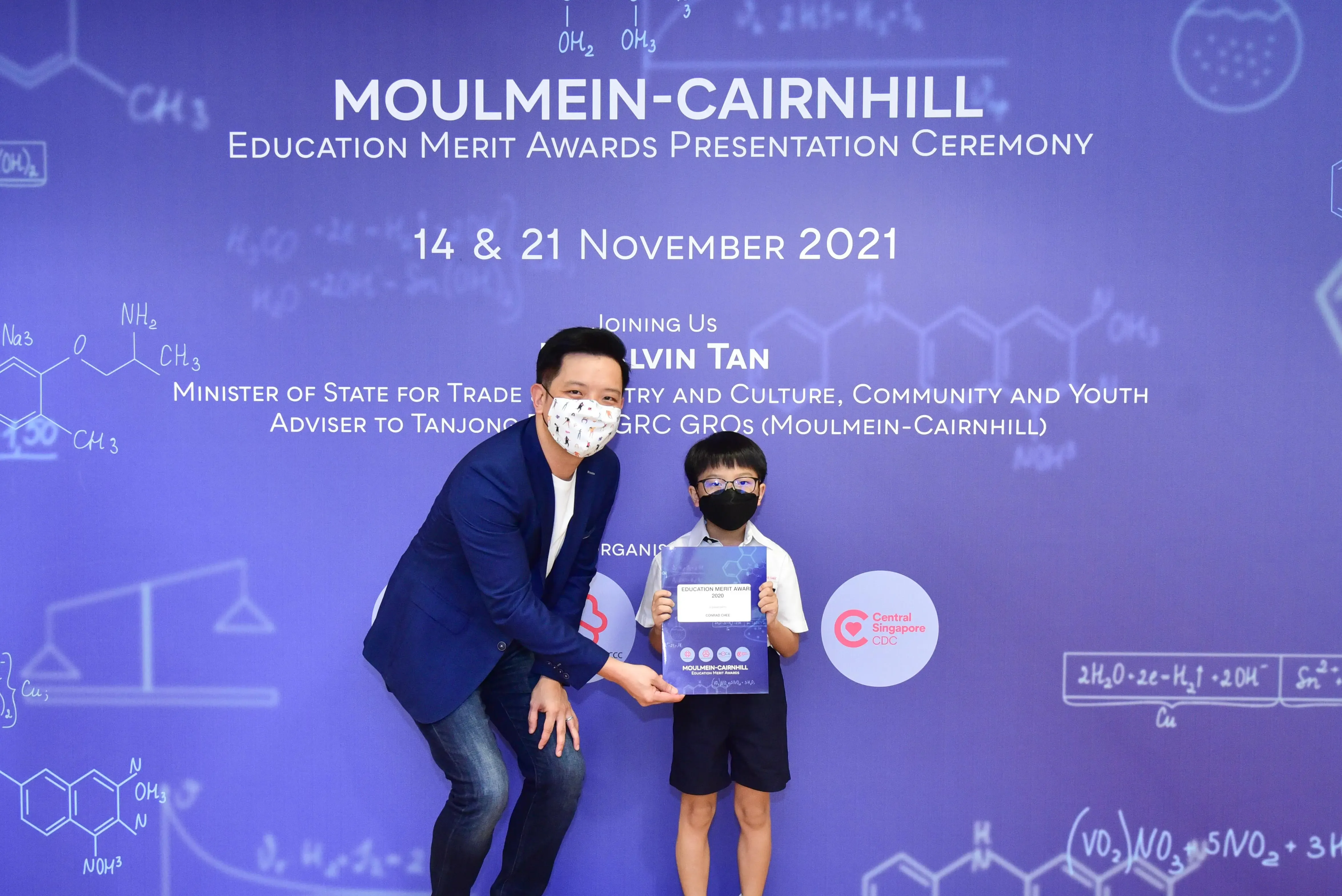 A masked man and young boy holding a certificate pose at the Moulmein-Cairnhill Education Merit Awards 2021 backdrop.