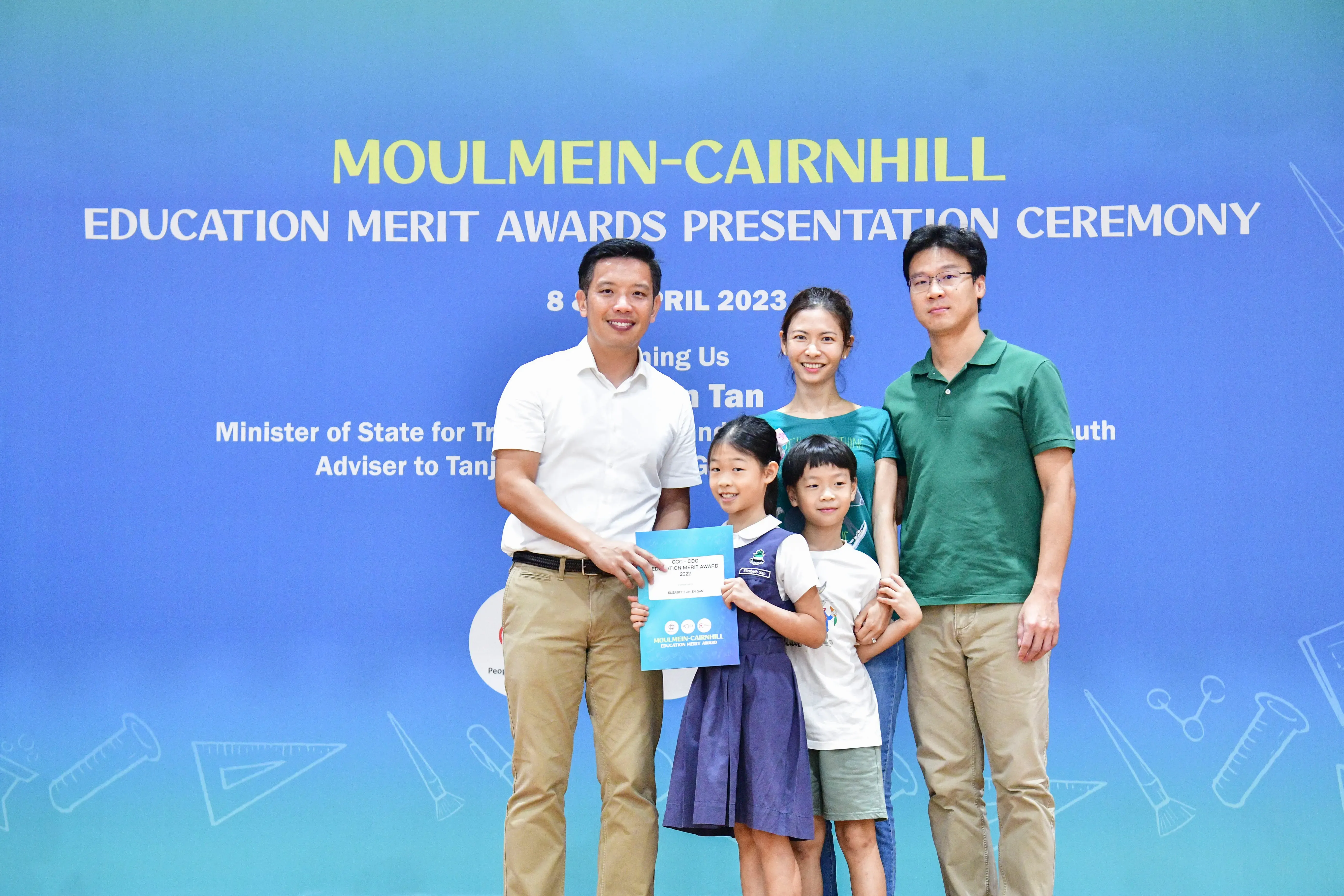 Five people pose before a blue Moulmein-Cairnhill Education Merit Awards Presentation Ceremony banner, girl holding certificate.
