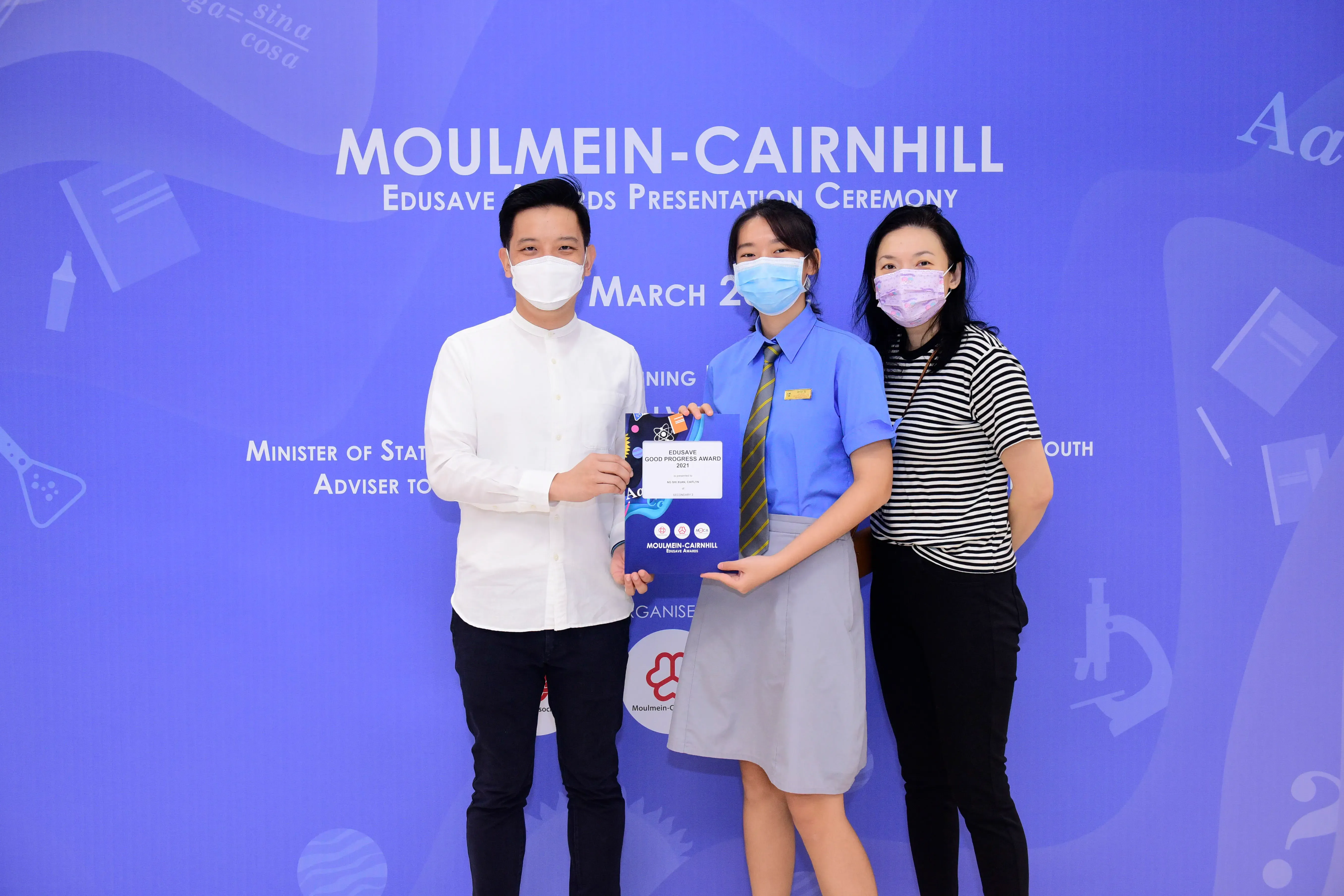 Three masked people pose at the Moulmein-Cairnhill Edusave Awards Presentation Ceremony, student holding an award folder.