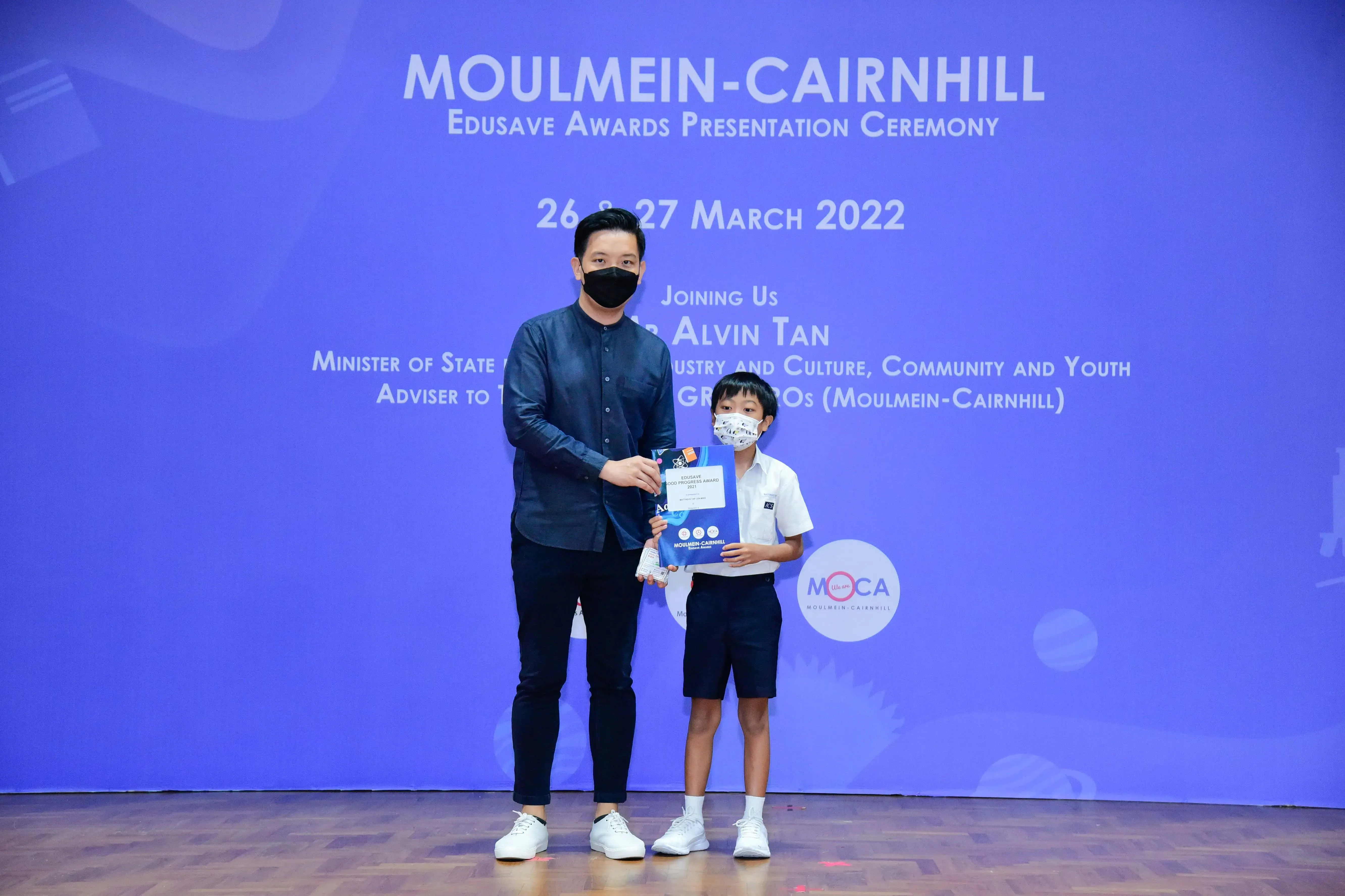 An adult and a young boy wearing masks hold an award certificate at the Moulmein-Cairnhill Edusave Awards Ceremony 2022.