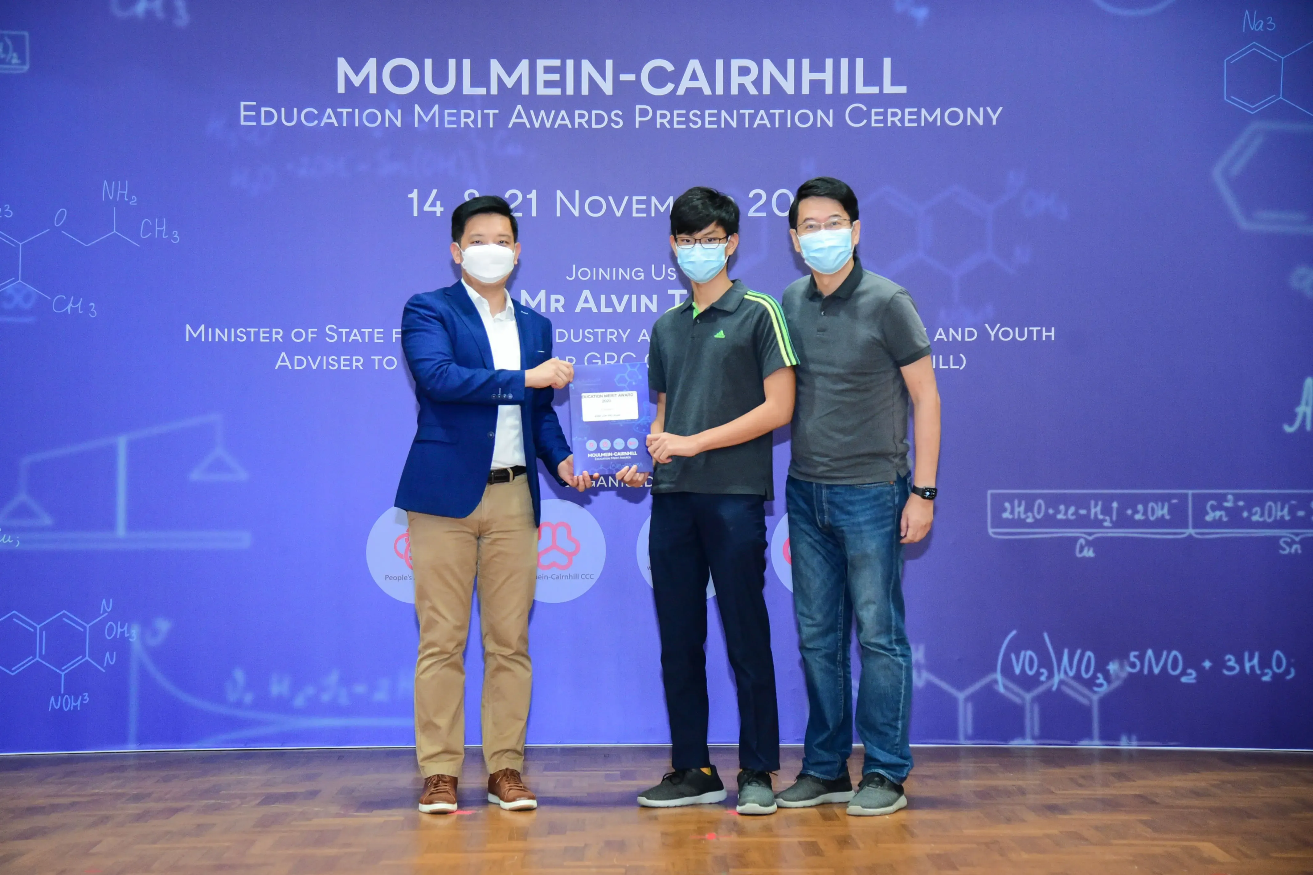 Three masked men stand before a Moulmein-Cairnhill Education Merit Awards Presentation Ceremony backdrop, one holding an award.