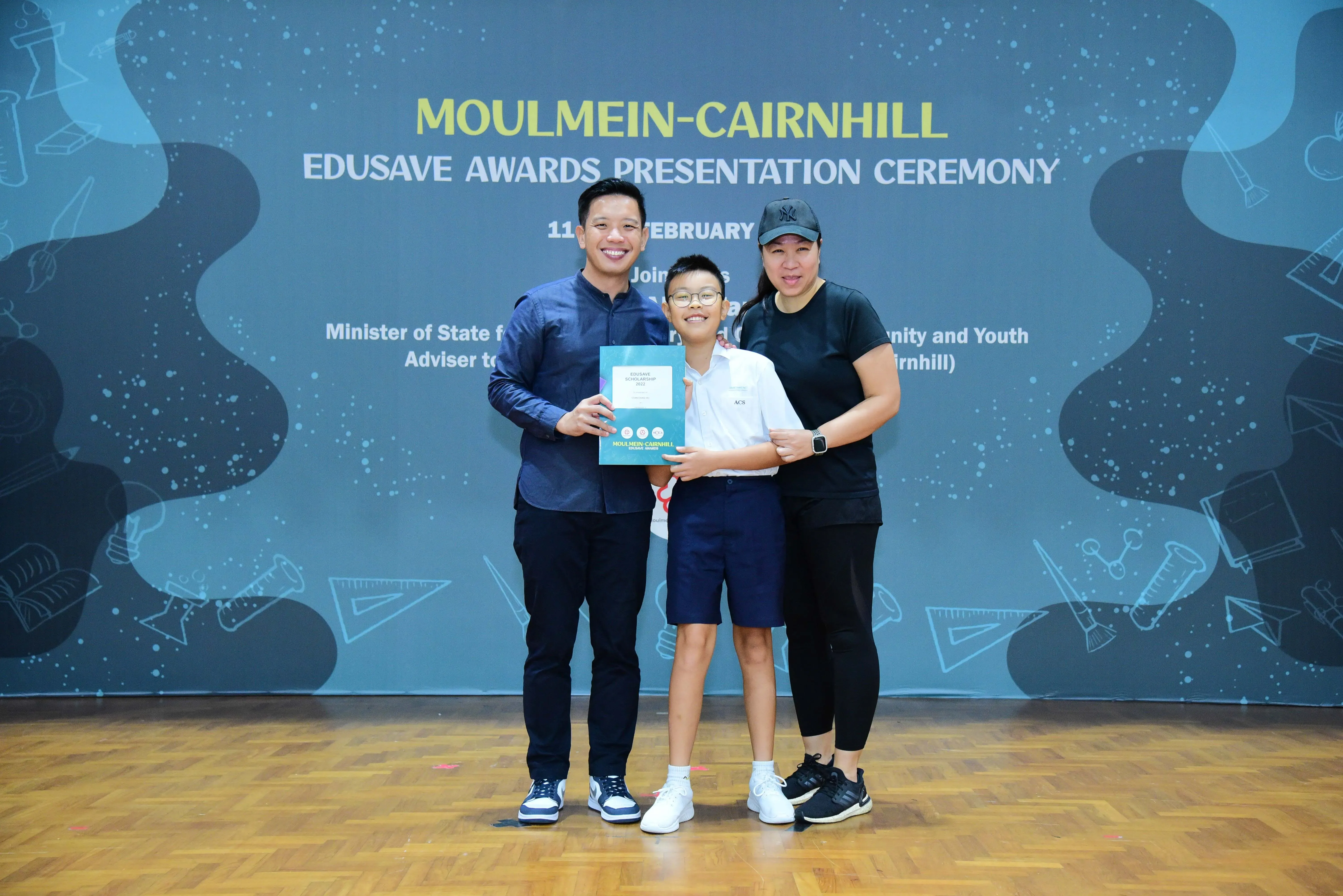 A boy in school uniform holds a certificate flanked by two adults at the Moulmein-Cairnhill Edusave Awards Ceremony.