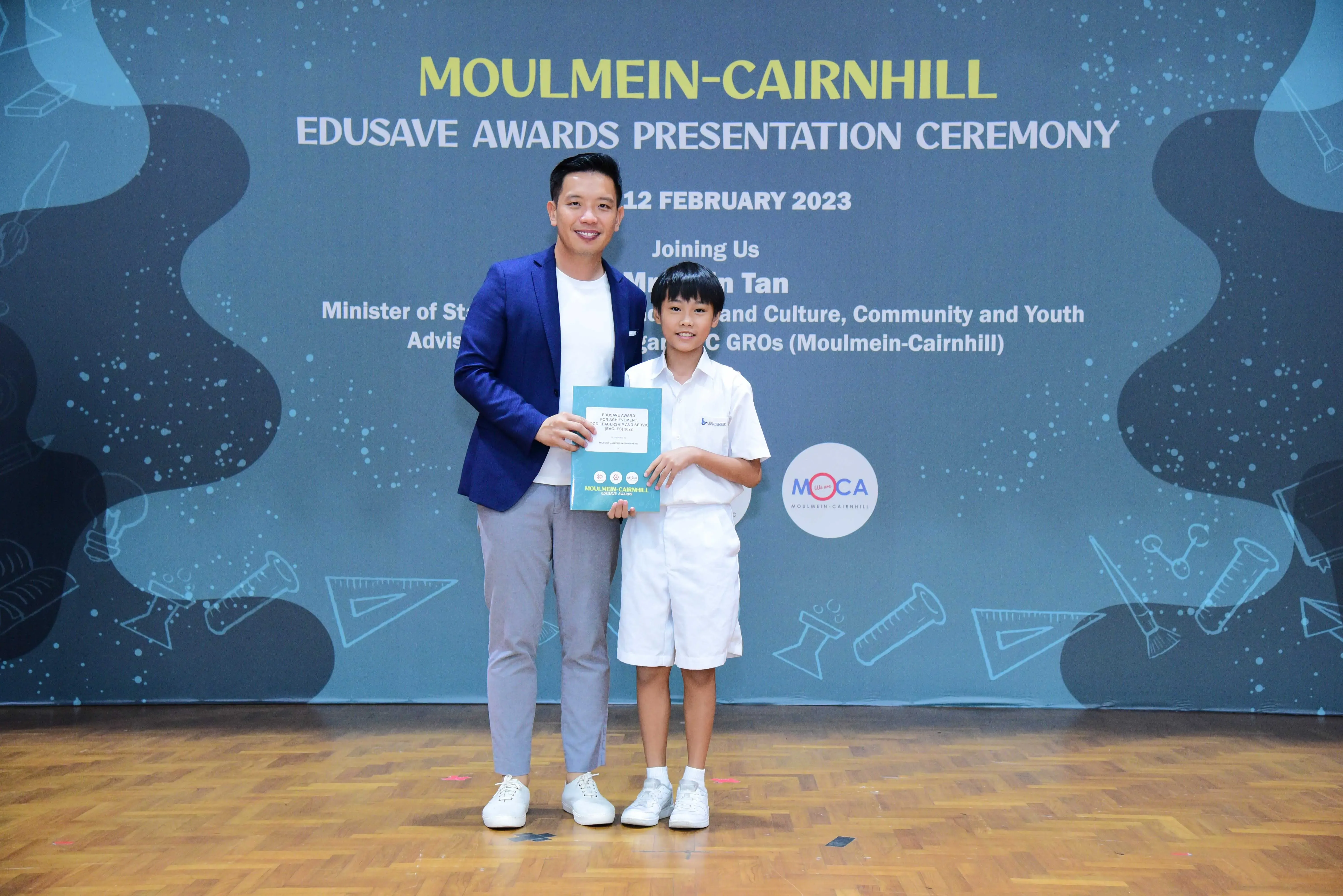 An adult in a blue blazer and a young boy in school uniform hold a certificate at the Moulmein-Cairnhill Edusave Awards ceremony.