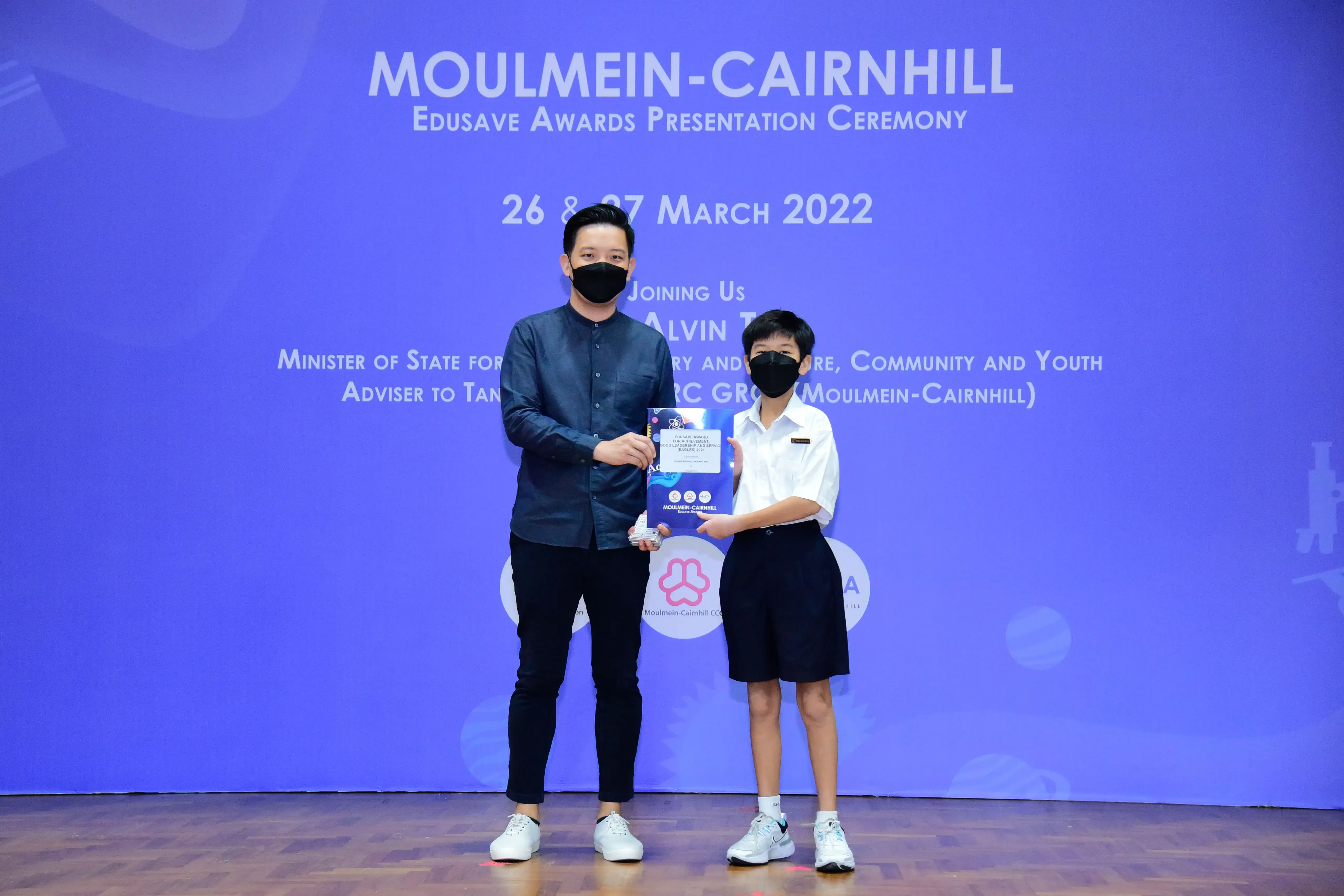 An adult and a child in masks hold a certificate on stage at the Moulmein-Cairnhill Edusave Awards Ceremony, March 2022.