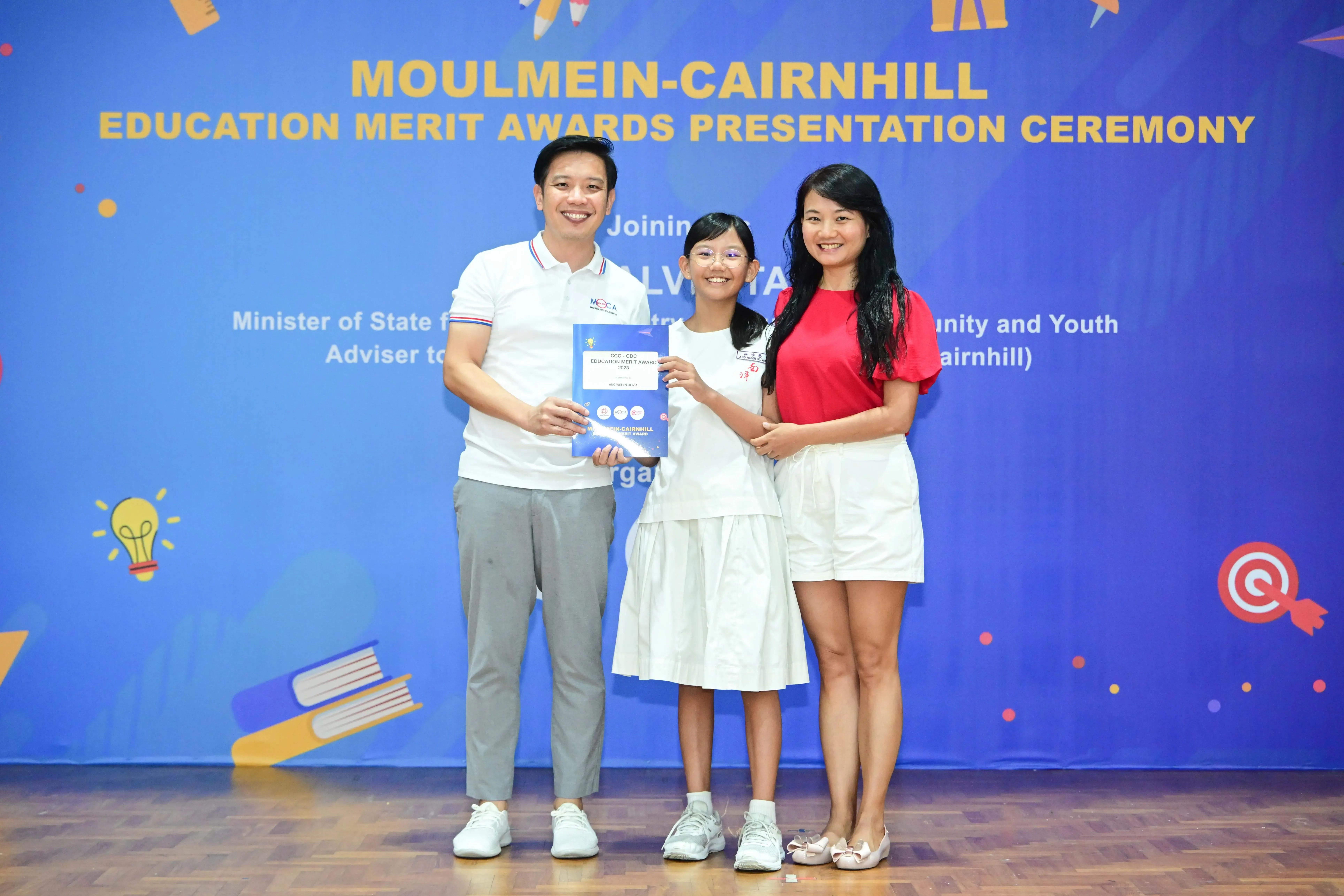 Three smiling people hold a certificate at the Moulmein-Cairnhill Education Merit Awards Presentation Ceremony.
