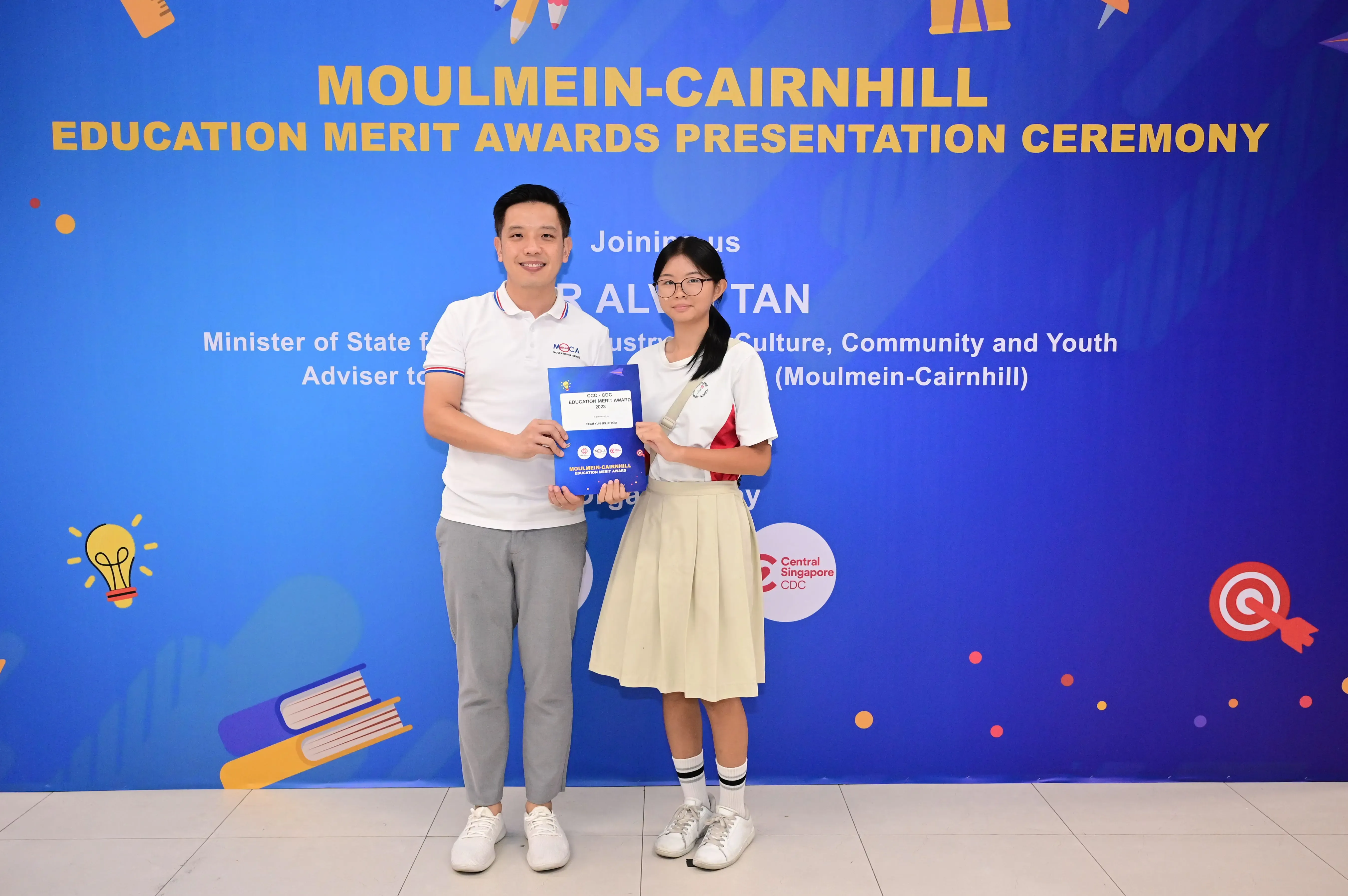 Two people hold an award booklet in front of a Moulmein-Cairnhill Education Merit Awards Presentation Ceremony banner.