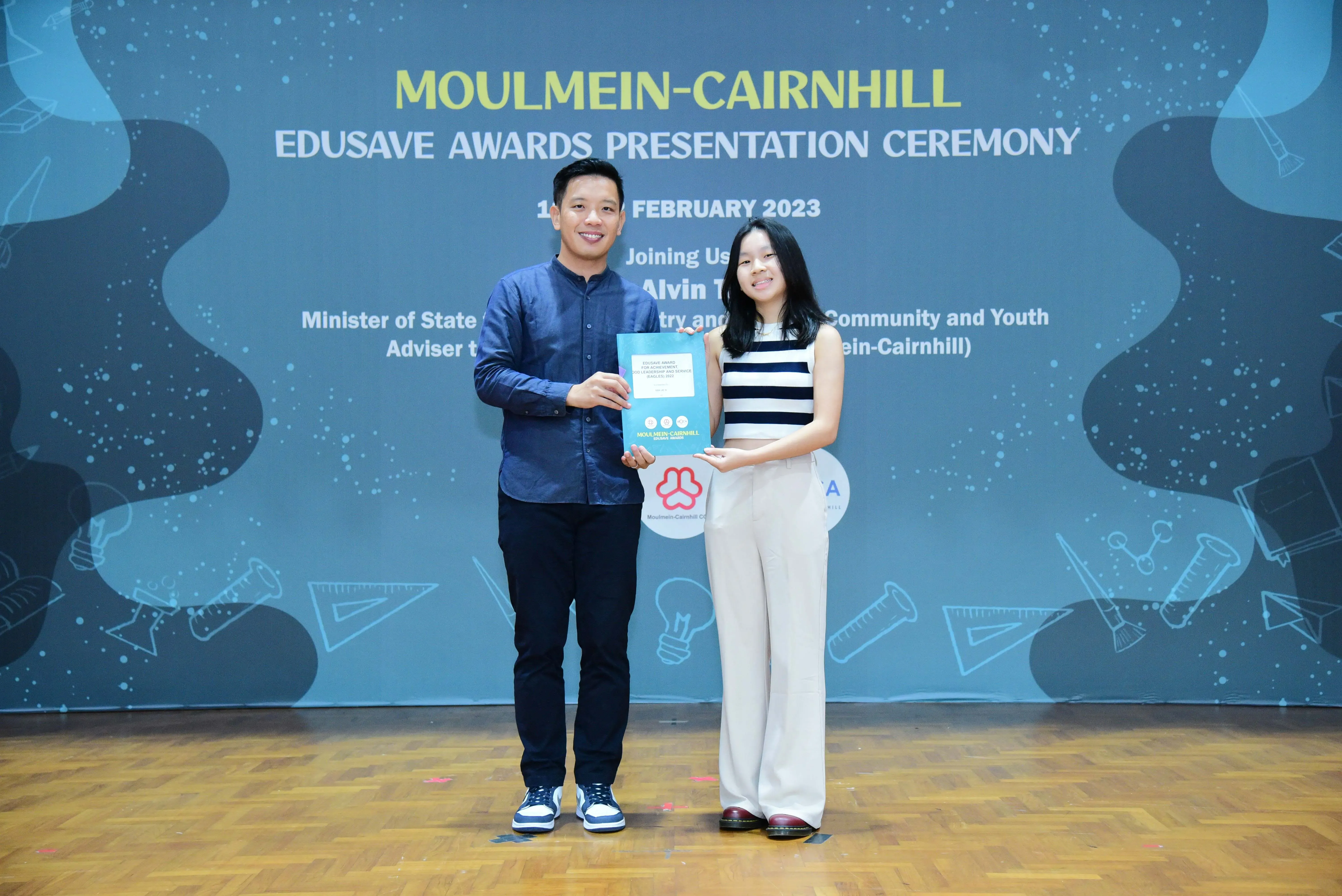 Two people stand on a stage holding a certificate at the Moulmein-Cairnhill Edusave Awards Ceremony, February 2023.