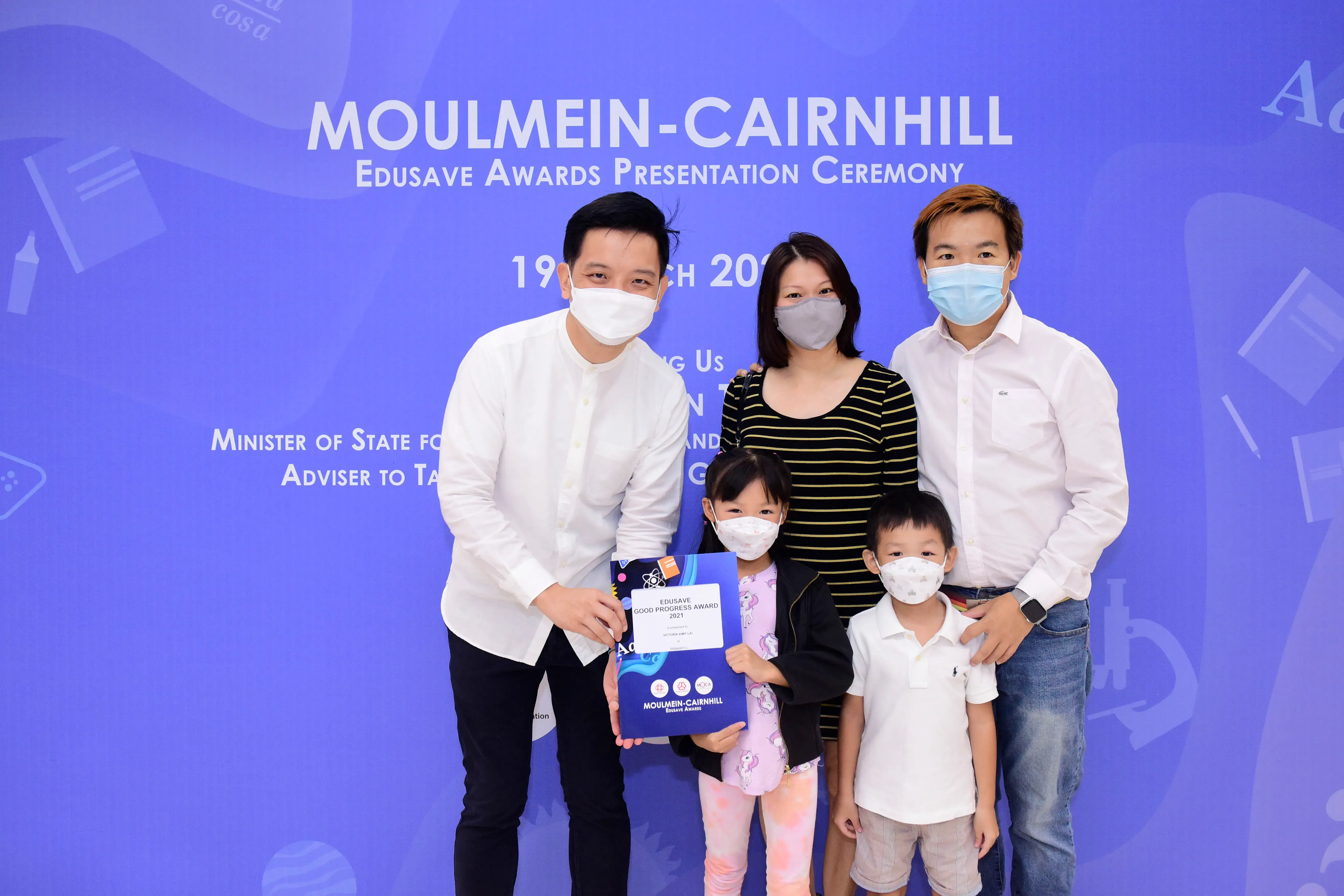 Five masked people pose at the Moulmein-Cairnhill Edusave Awards Presentation Ceremony, with a child holding an award.