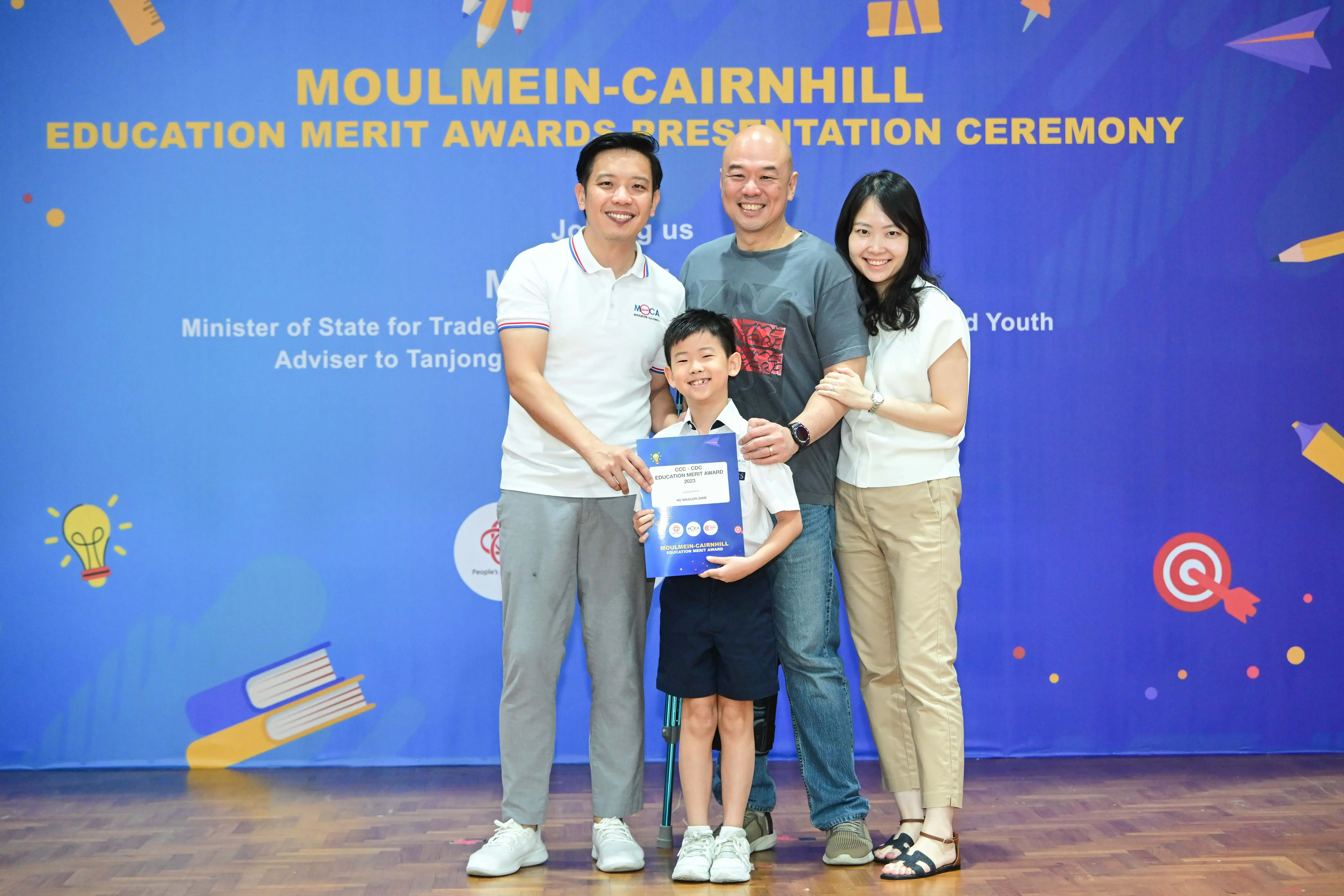 Three adults and a young boy holding a certificate pose before a Moulmein-Cairnhill Education Merit Awards banner.