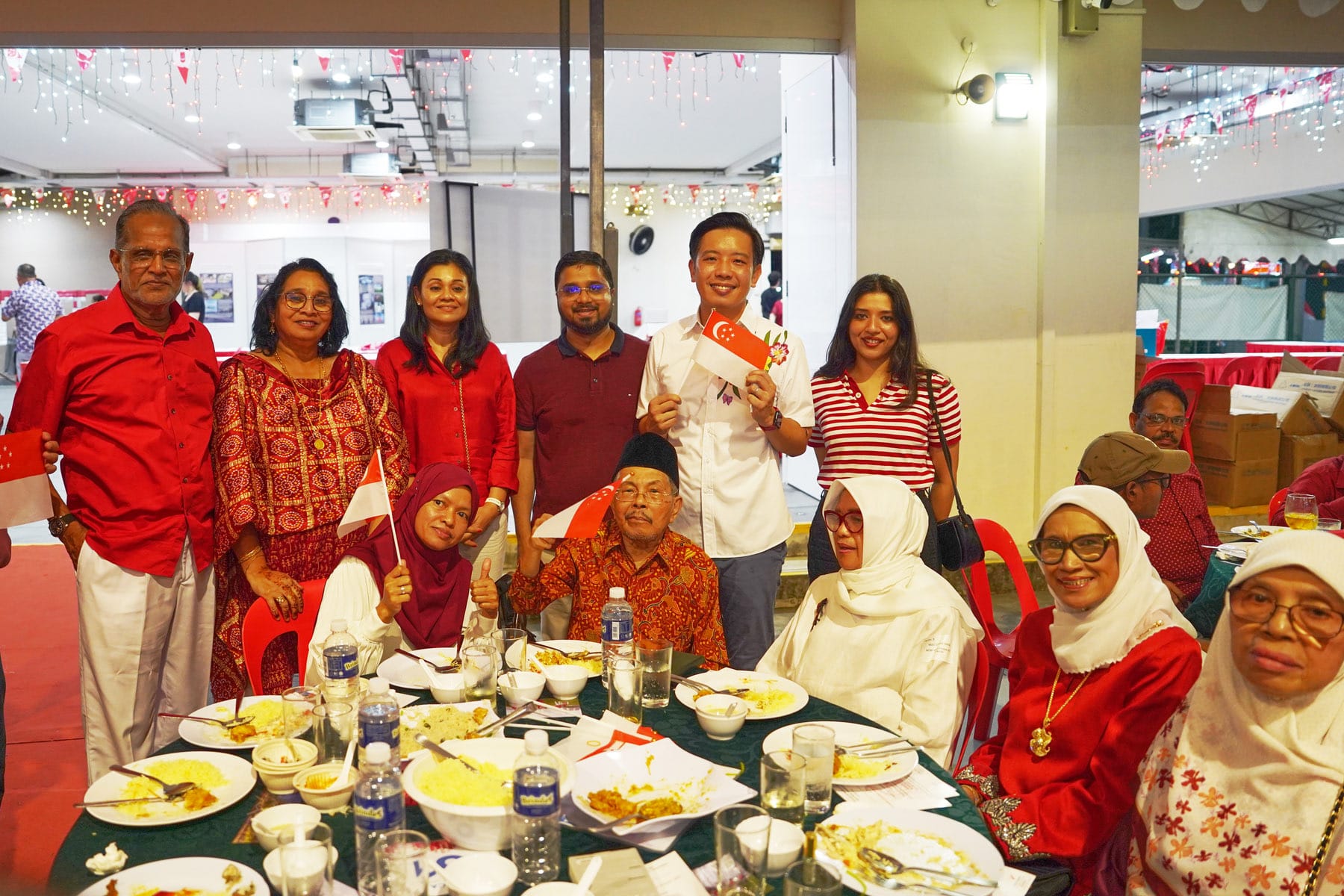 Group of people in red and white attire posing together at a dinner table with food and small Singapore flags.