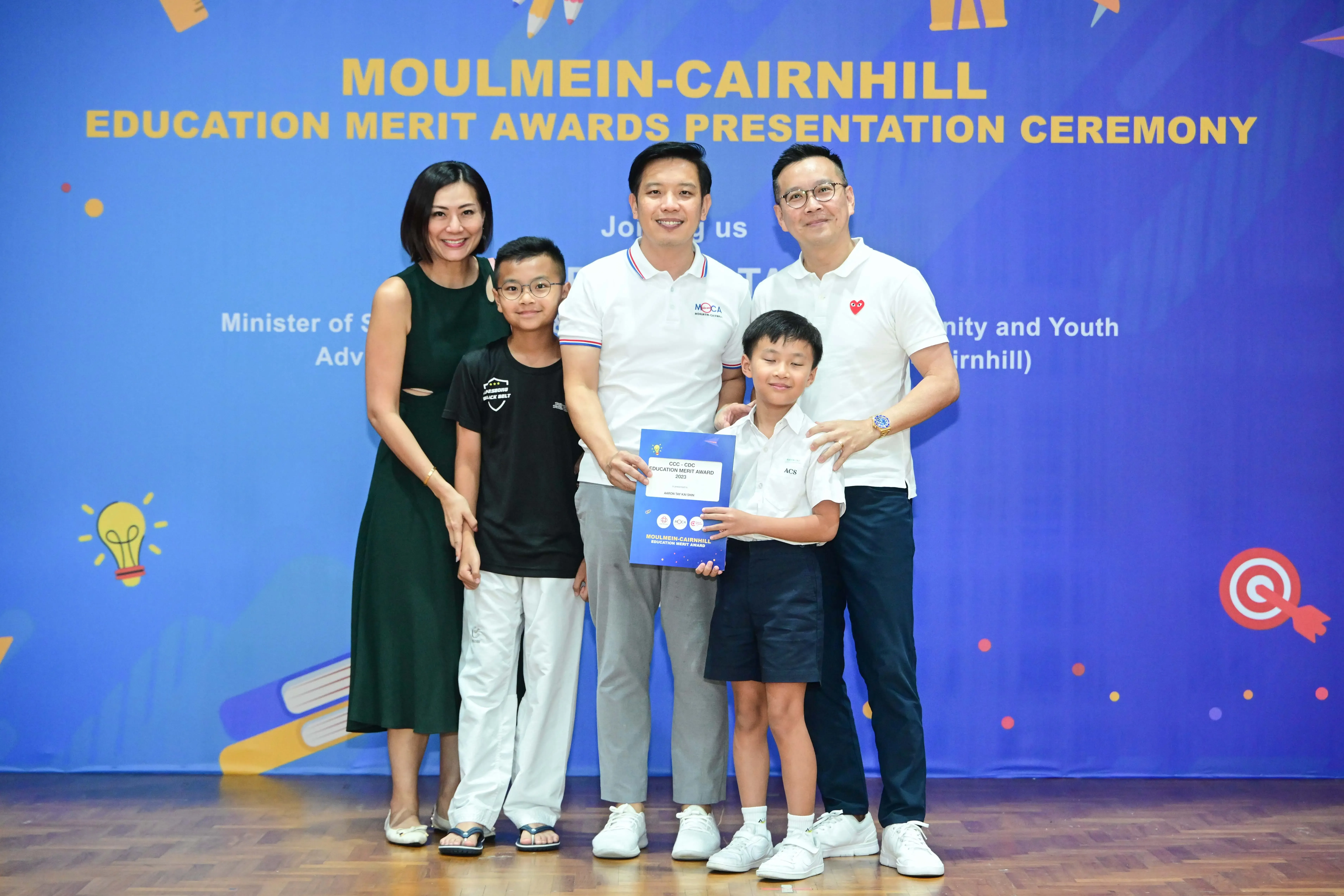 Five people pose at the Moulmein-Cairnhill Education Merit Awards Presentation Ceremony, with a boy holding a blue award certificate.