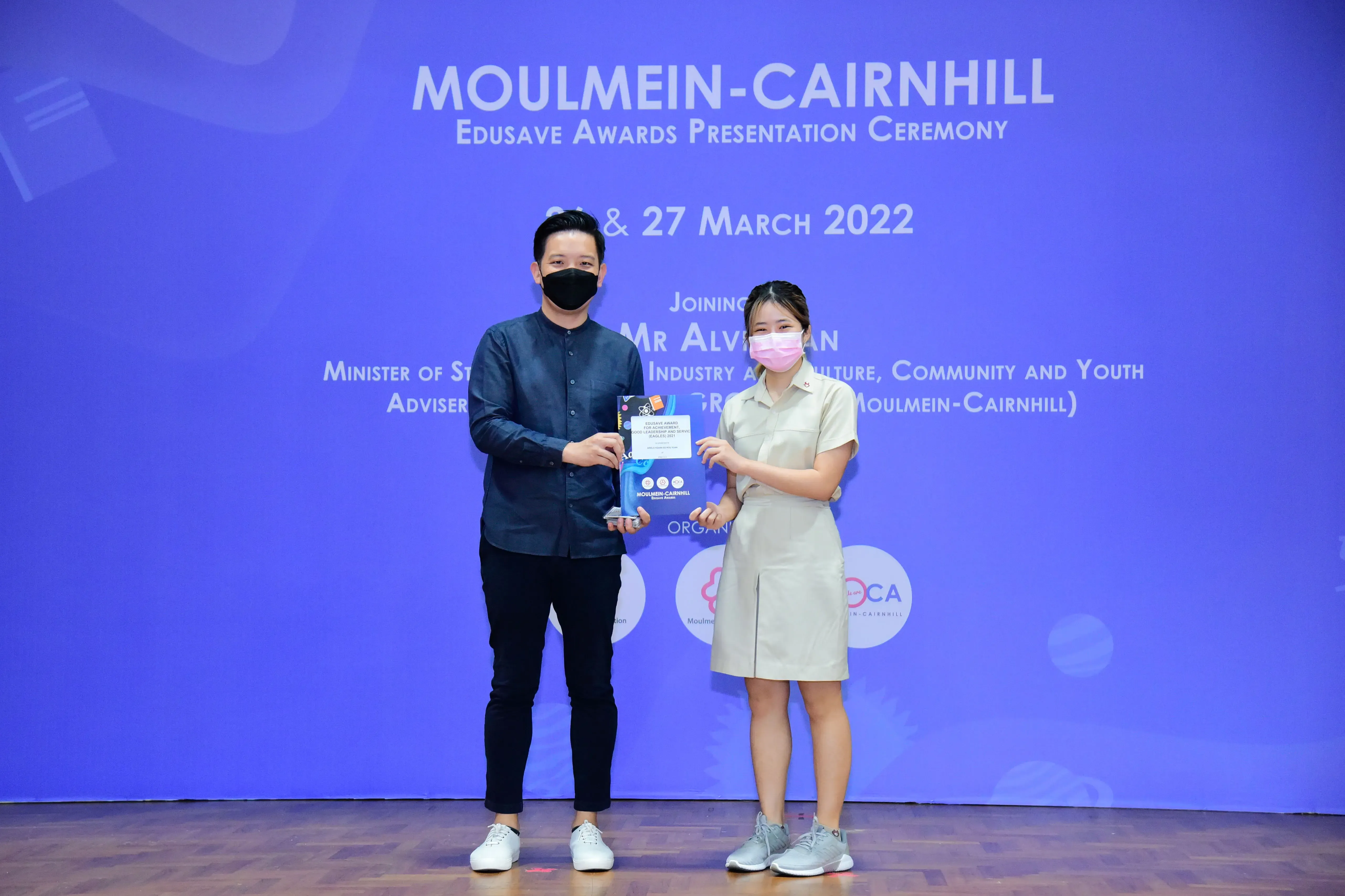 Two masked people hold an award at the Moulmein-Cairnhill Edusave Awards Presentation Ceremony, March 2022.