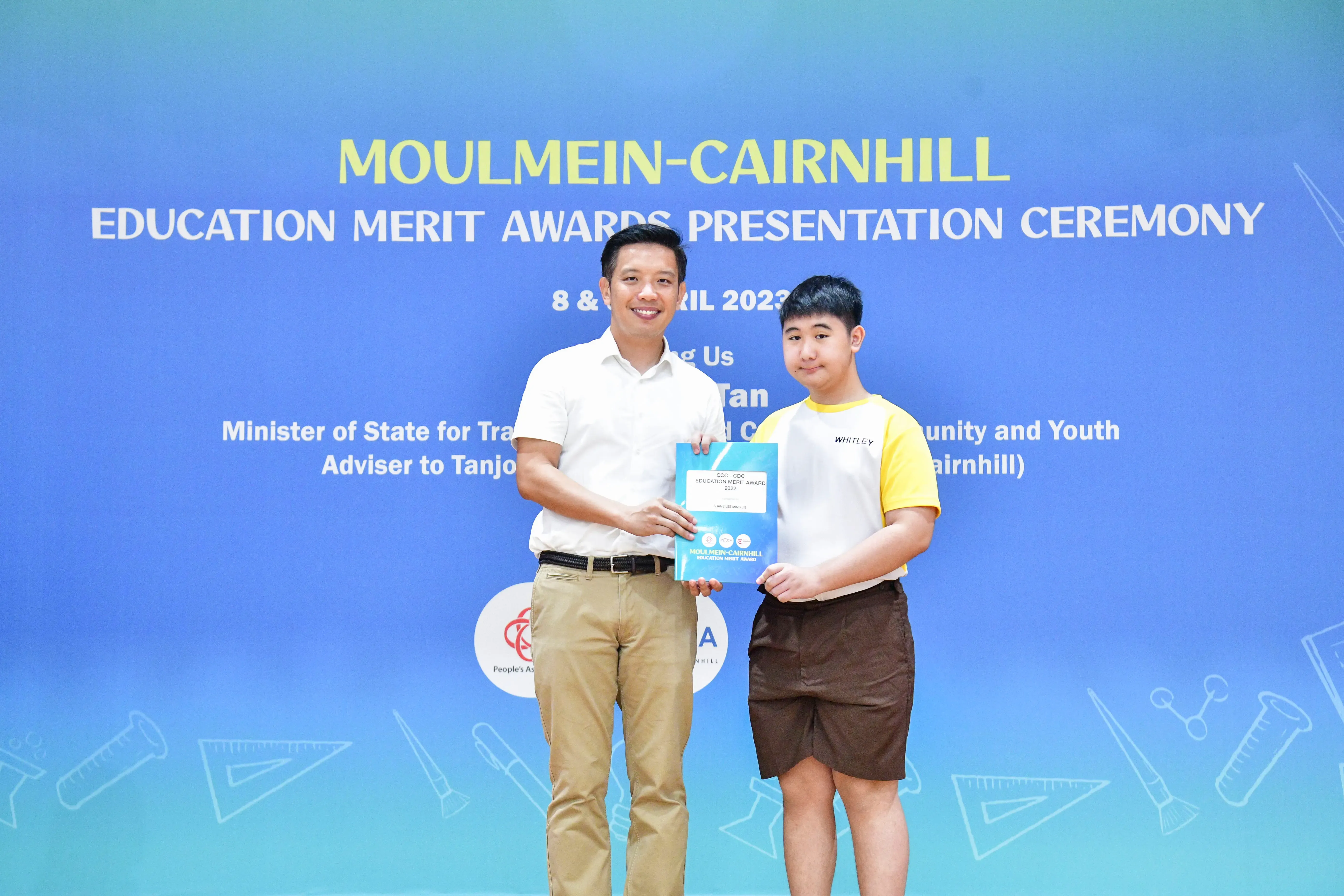 Two people hold an award certificate in front of a blue Moulmein-Cairnhill Education Merit Awards Ceremony backdrop.