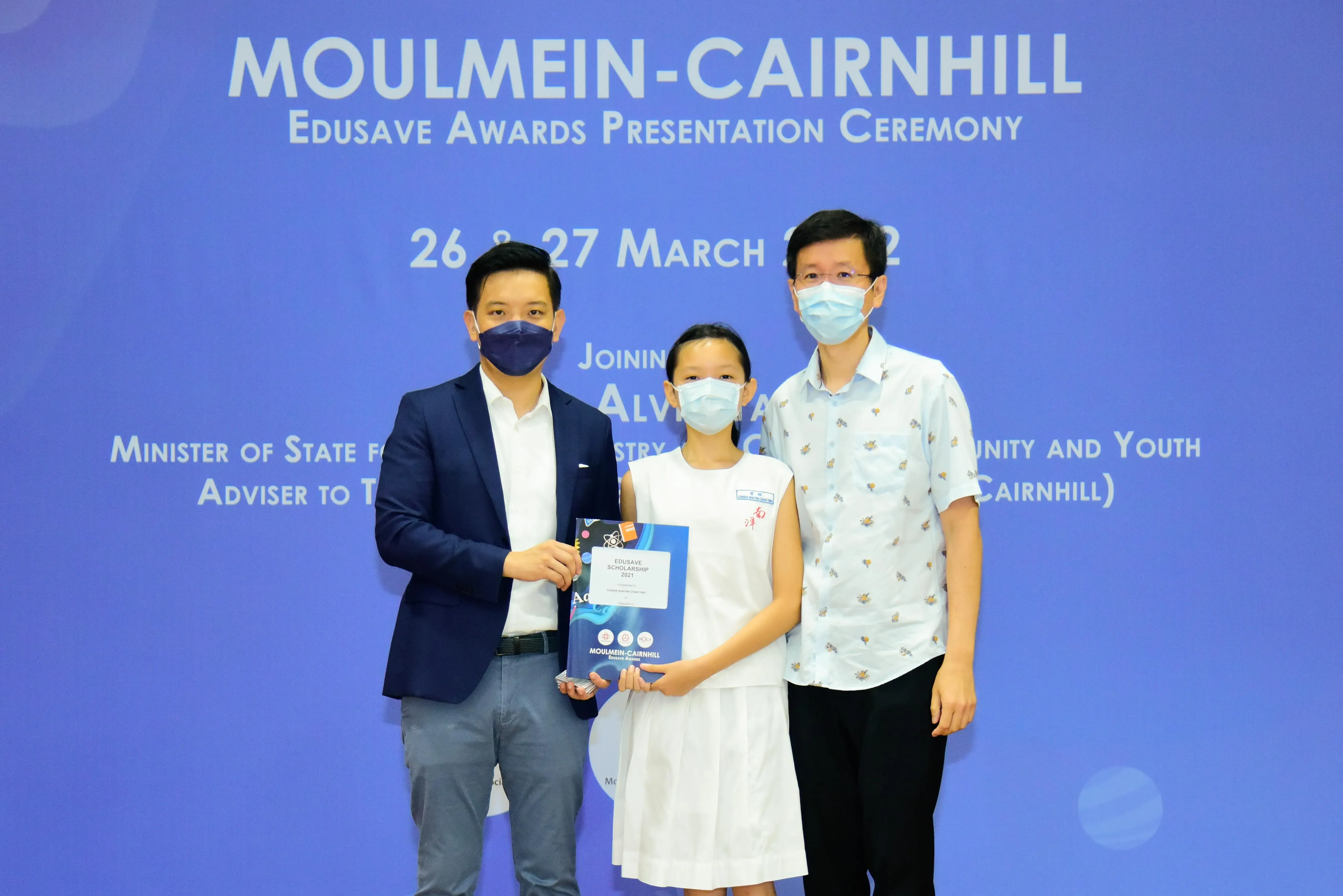 Three masked people pose at the Moulmein-Cairnhill Edusave Awards Presentation Ceremony, with a girl holding an award folder.