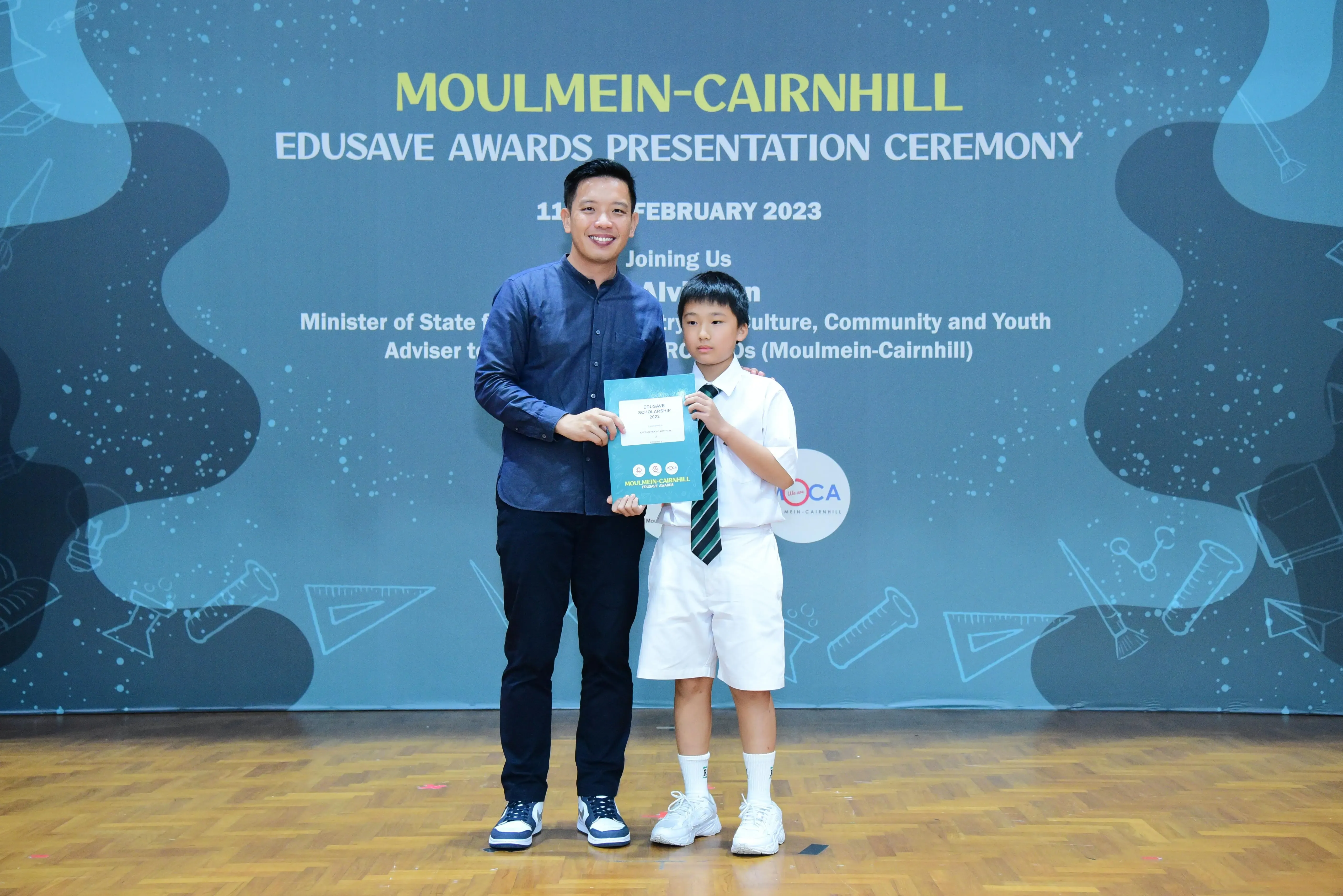 An adult and a young student hold a certificate on stage at the Moulmein-Cairnhill Edusave Awards Ceremony 2023.