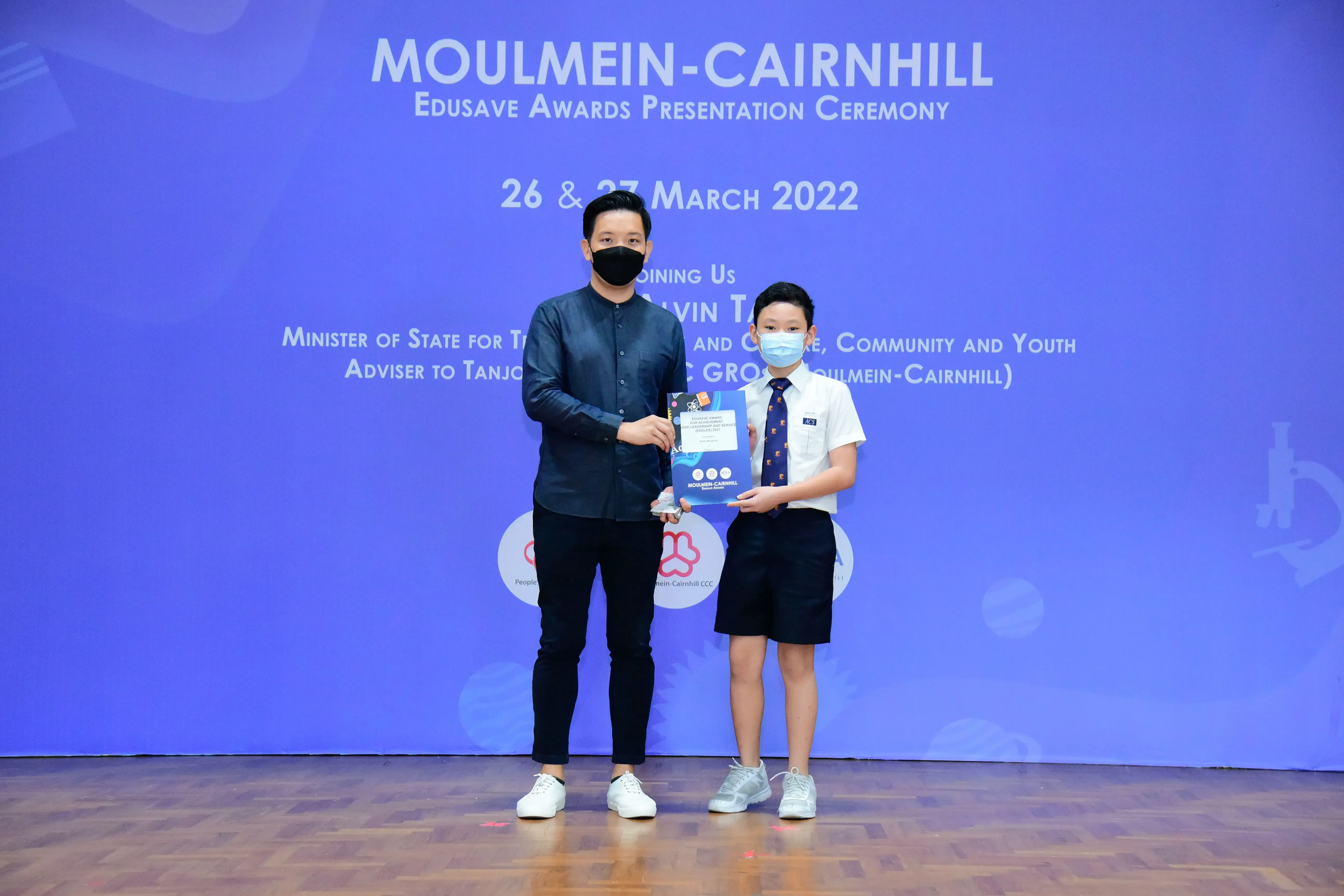 An adult and a young student hold an award together at the Moulmein-Cairnhill Edusave Awards Ceremony, March 2022.