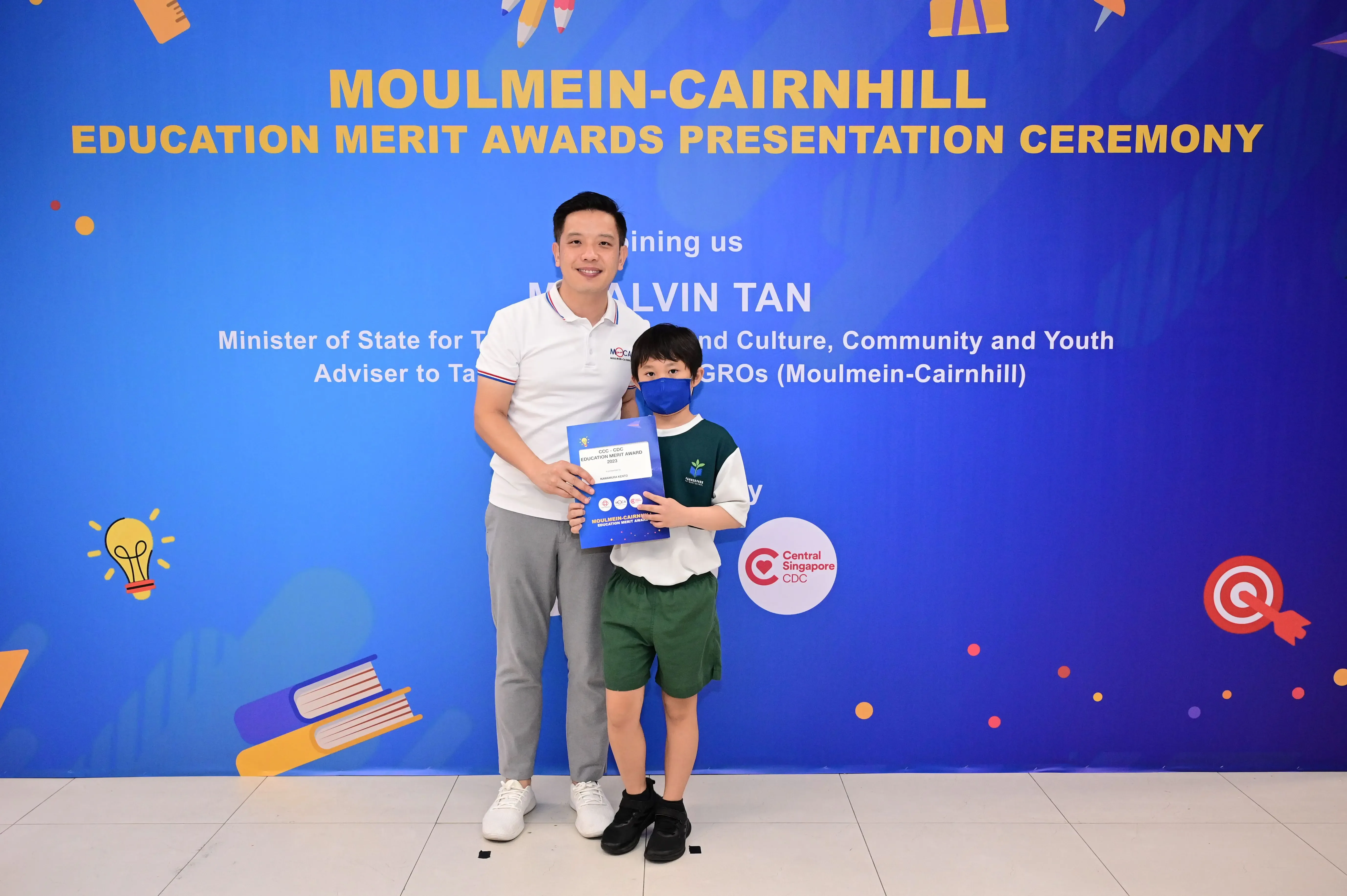 An adult and a masked child hold an award certificate in front of a Moulmein-Cairnhill Education Merit Awards banner.