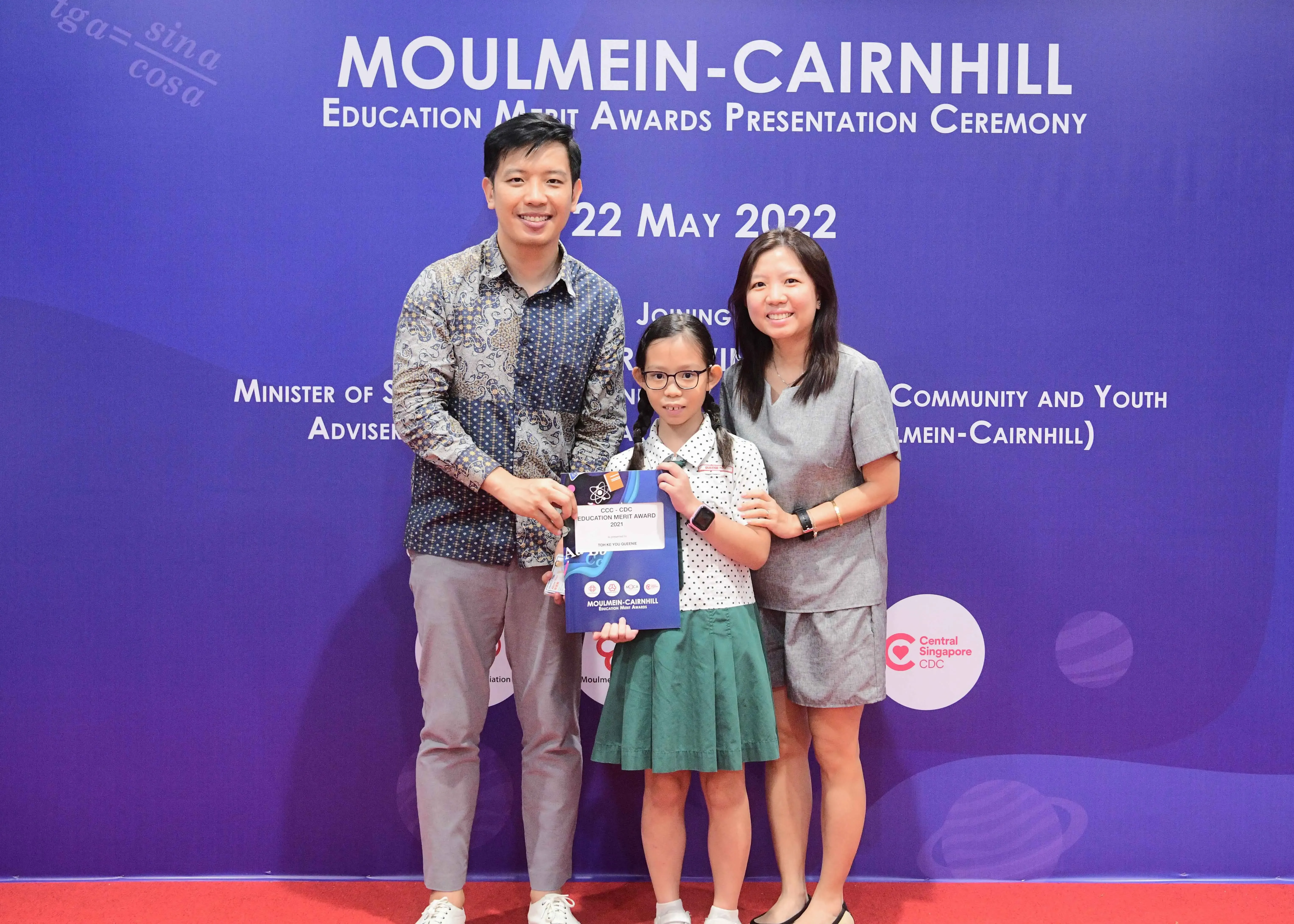 Two adults and a young girl holding an award certificate pose at the Moulmein-Cairnhill Education Merit Awards ceremony on 22 May 2022.