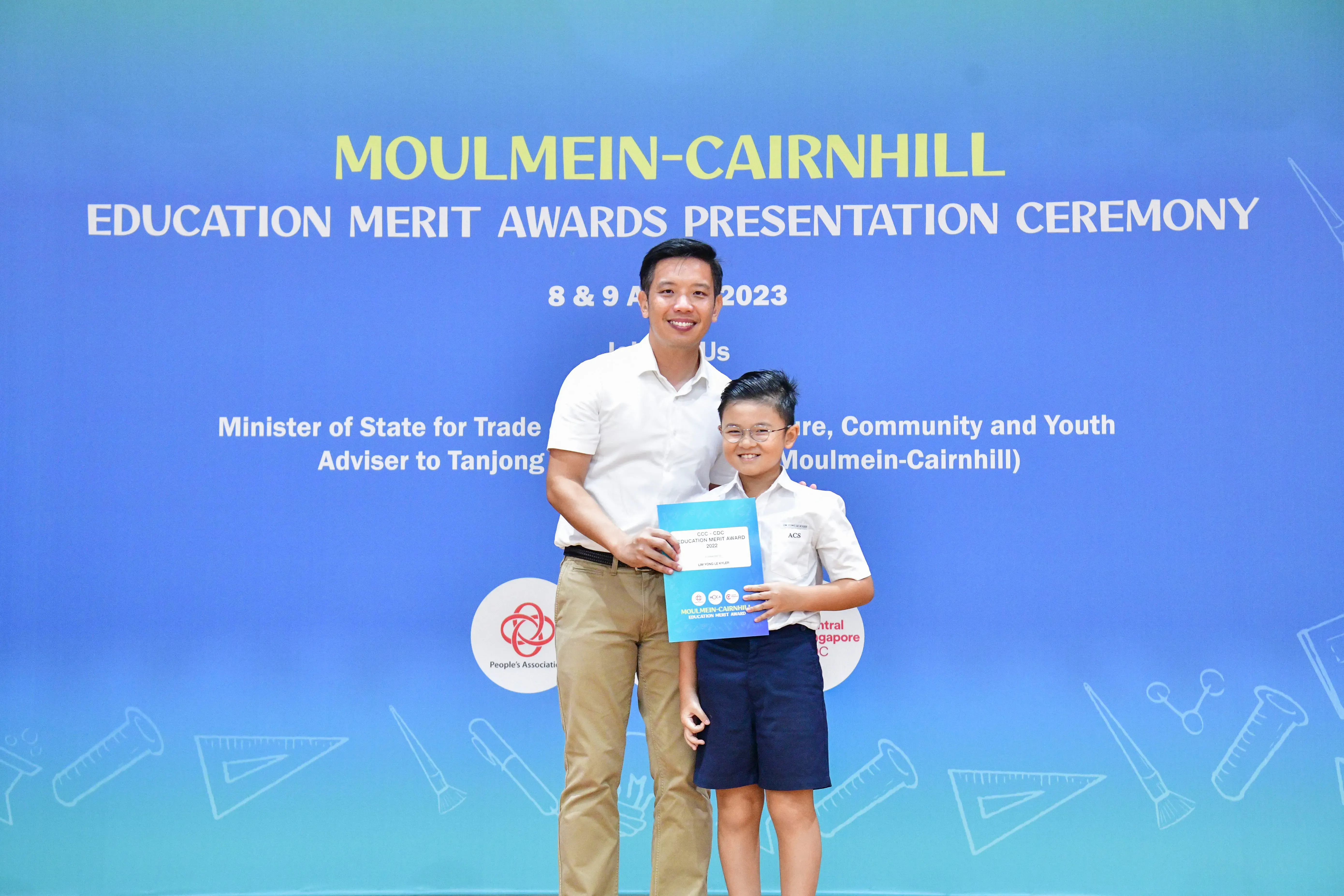 A man and a young boy holding a certificate pose before a Moulmein-Cairnhill Education Merit Awards 2023 banner.