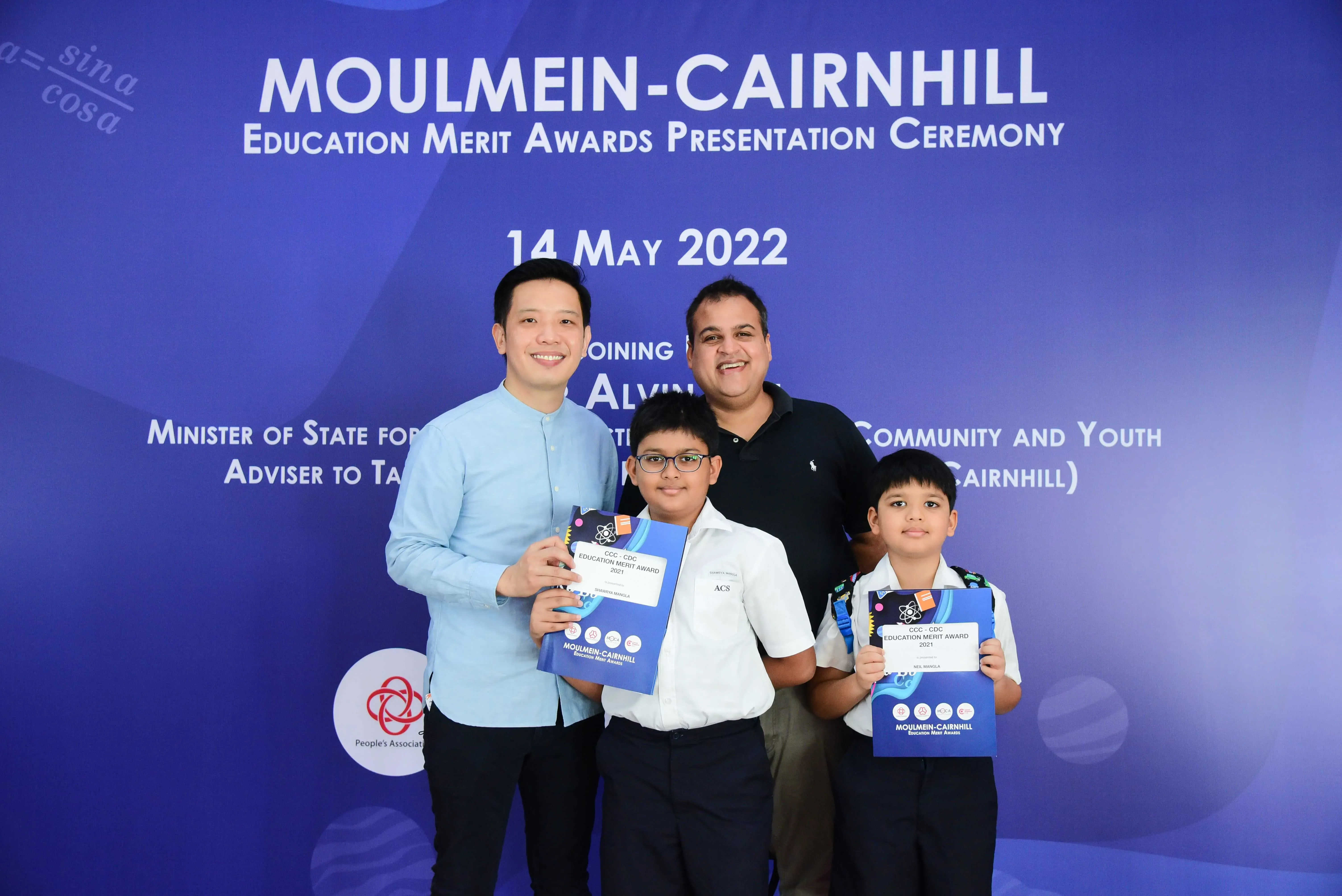 Two men and two young boys pose holding merit award folders at the Moulmein-Cairnhill Education Merit Awards ceremony, May 2022.