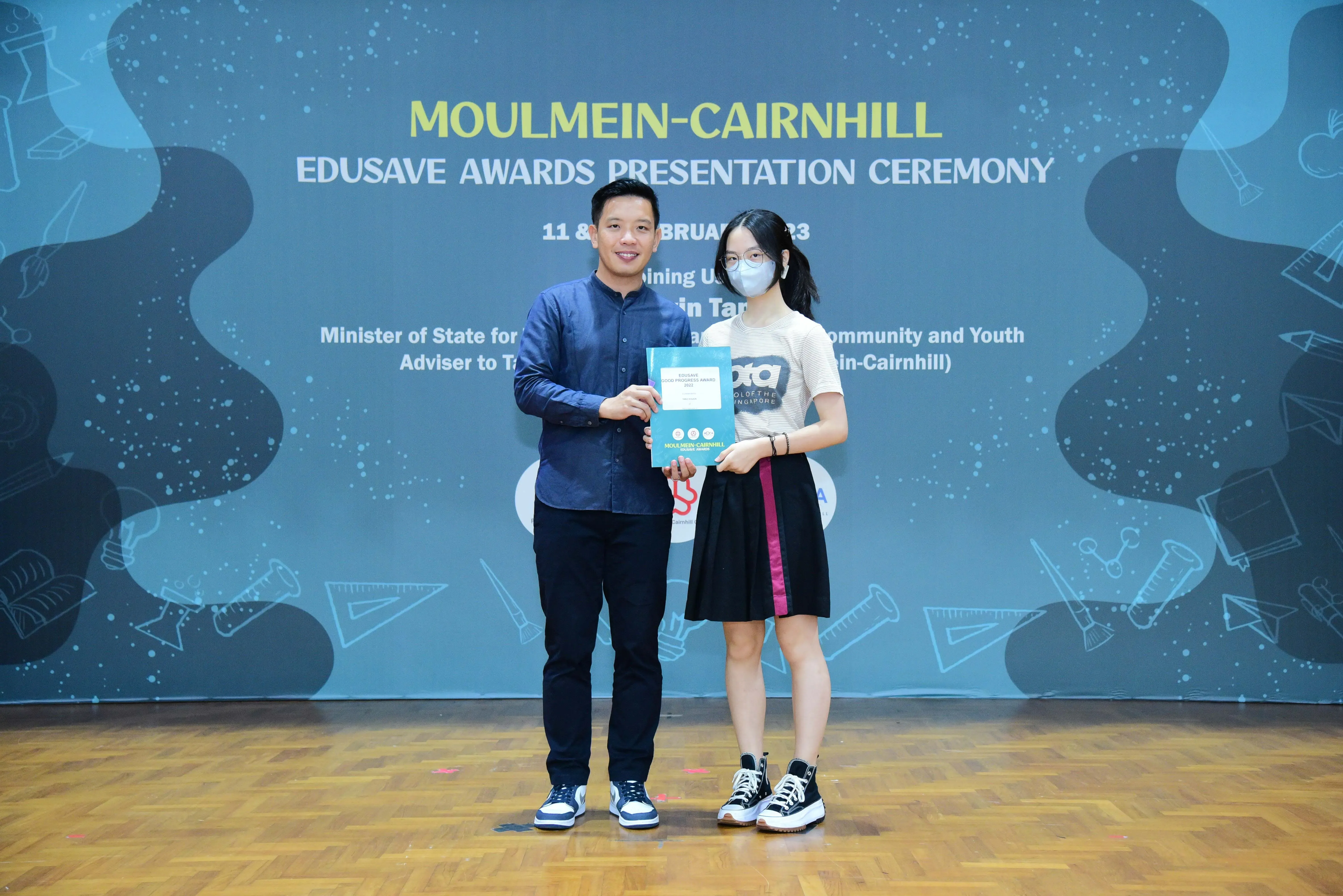 Two people hold a certificate together at the Moulmein-Cairnhill Edusave Awards Presentation Ceremony backdrop.