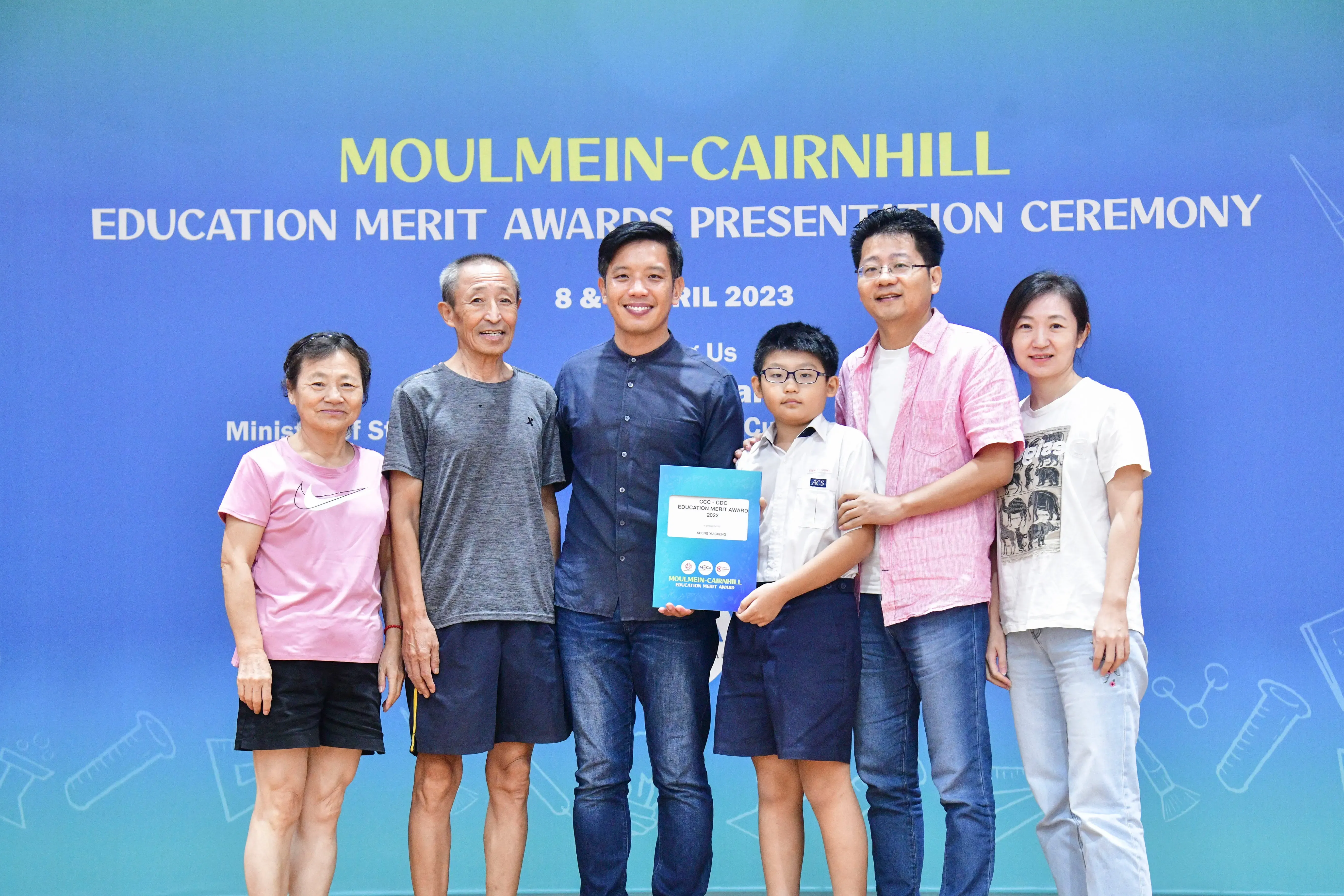 Six people pose together at the Moulmein-Cairnhill Education Merit Awards Presentation Ceremony, holding a blue certificate.