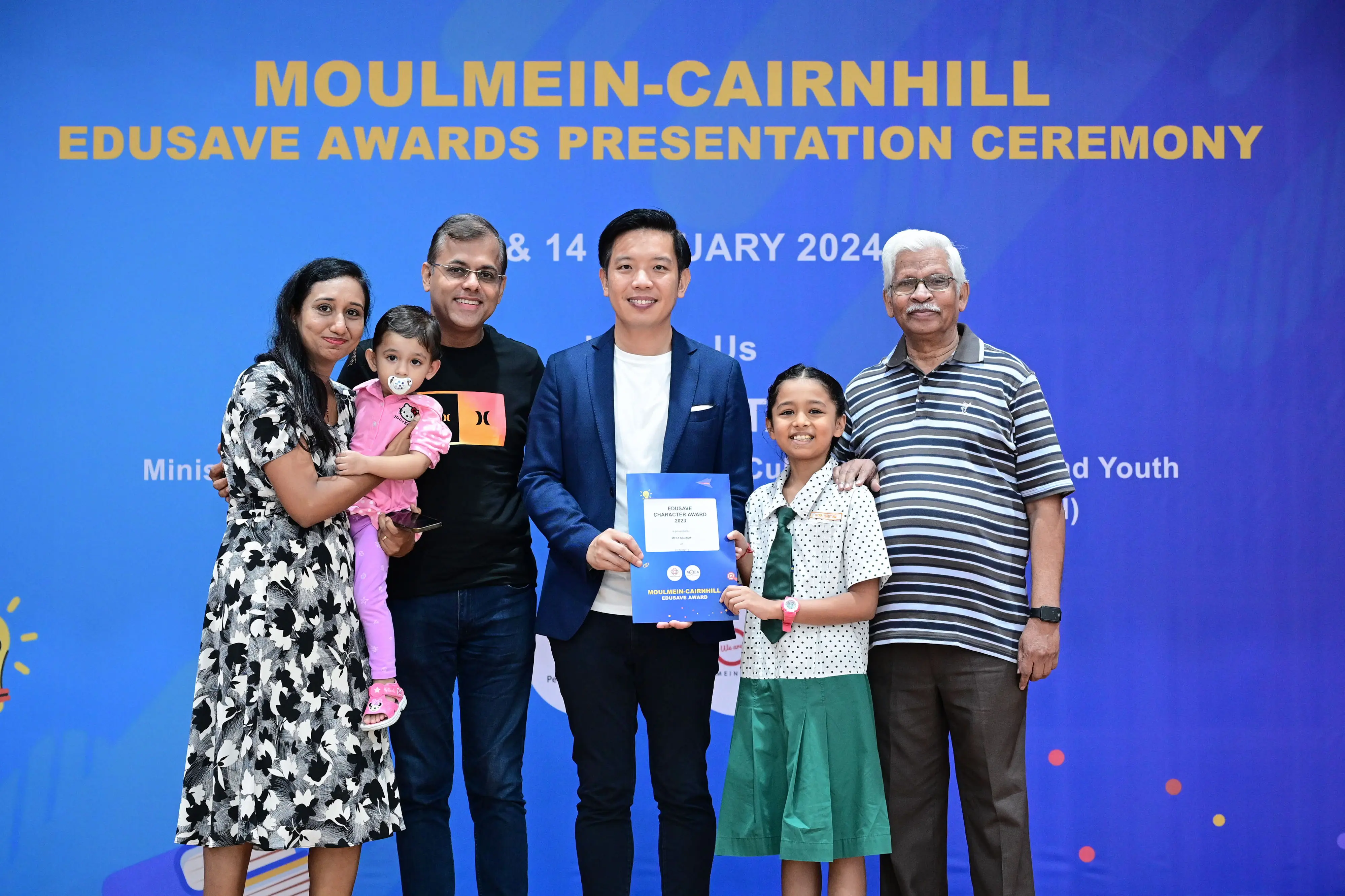 Six people pose at the Moulmein-Cairnhill Edusave Awards Presentation Ceremony 2024, with a girl holding a certificate.