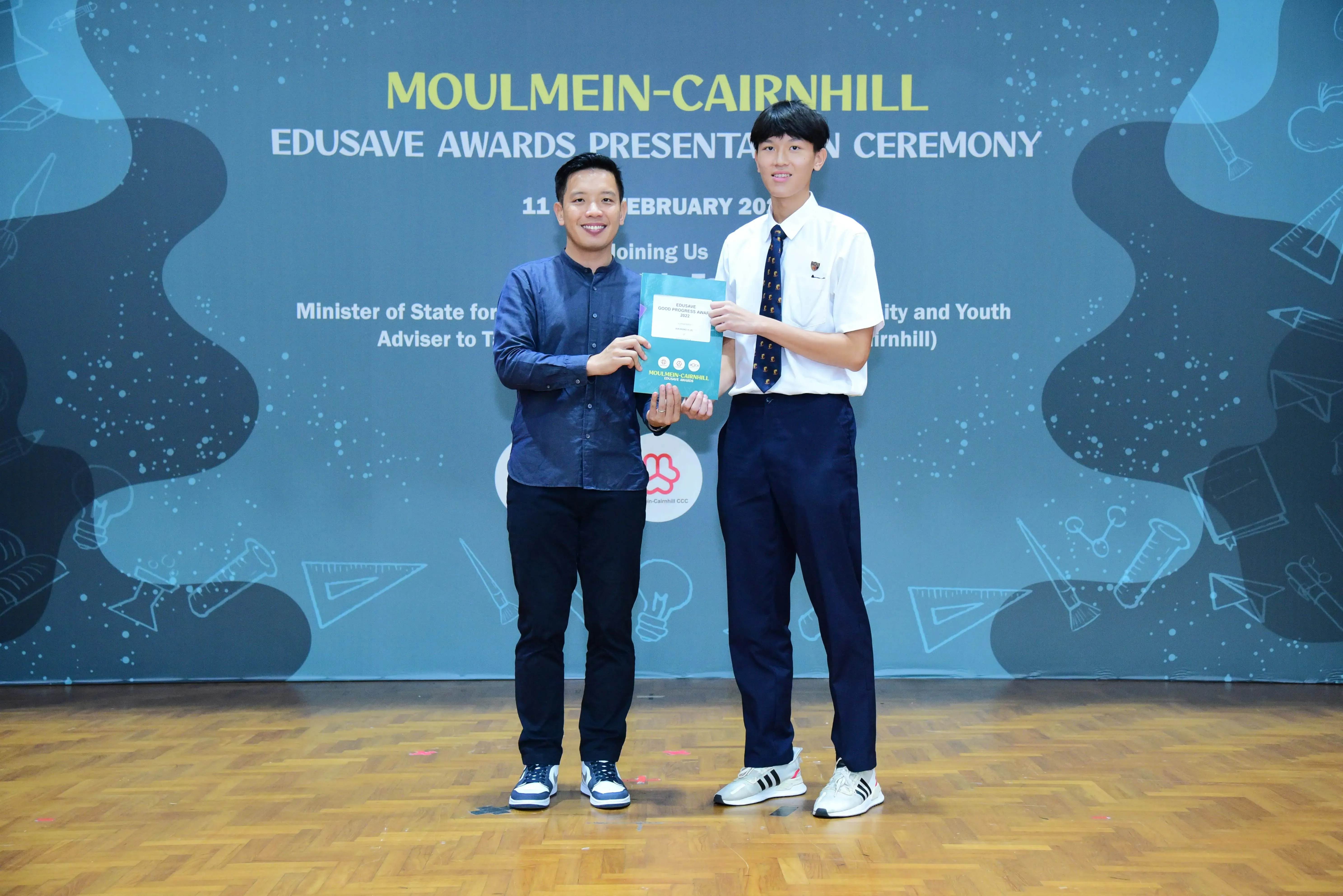 Two men hold a certificate together at the Moulmein-Cairnhill Edusave Awards Presentation Ceremony backdrop.