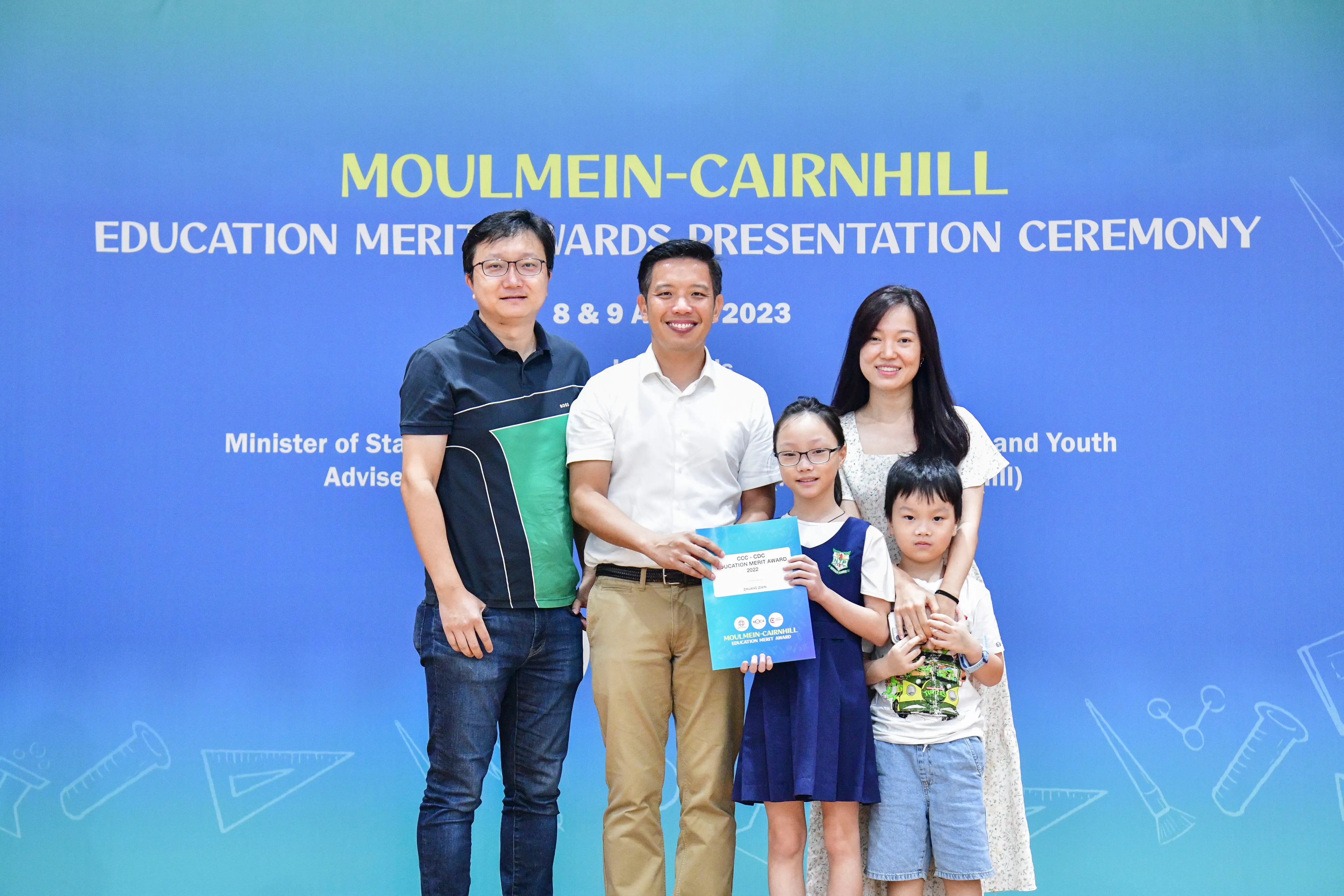 Five people pose before a Moulmein-Cairnhill Education Merit Awards 2023 banner, with a girl holding a certificate.