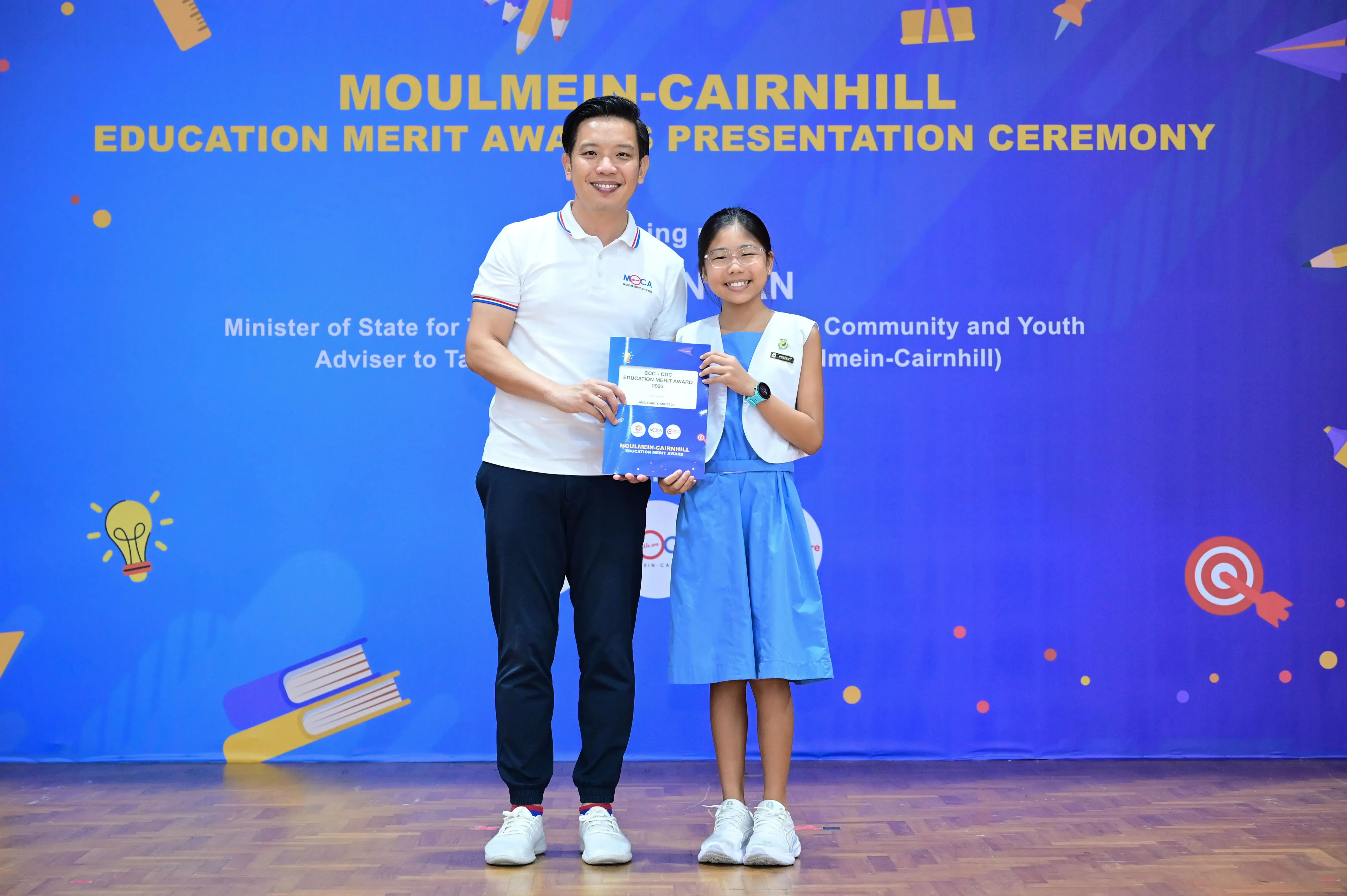 A man and a young girl hold an award certificate in front of a Moulmein-Cairnhill Education Merit Award banner.