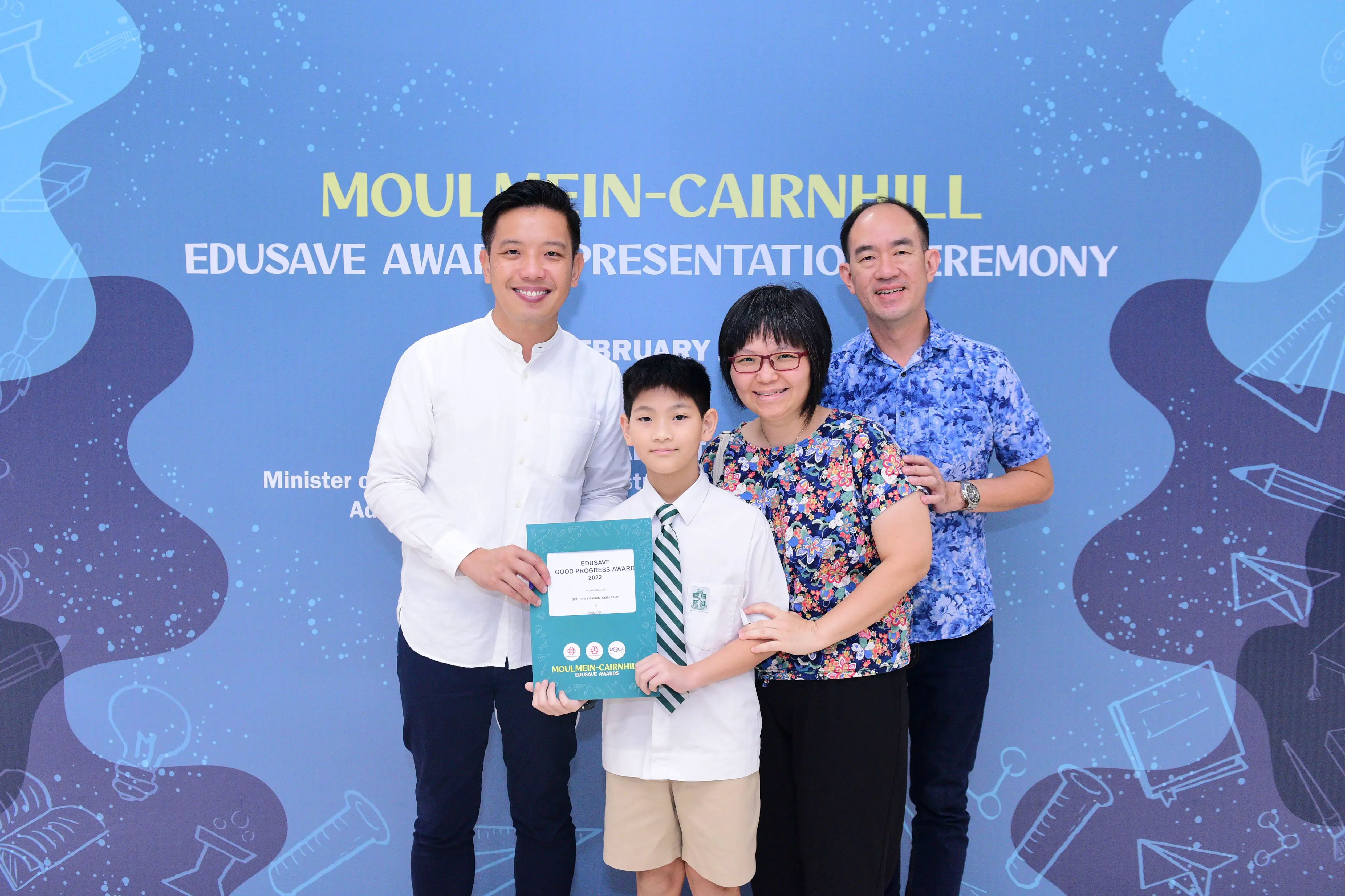 A boy in school uniform holds an Edusave award certificate, flanked by three smiling adults at a Moulmein-Cairnhill ceremony.