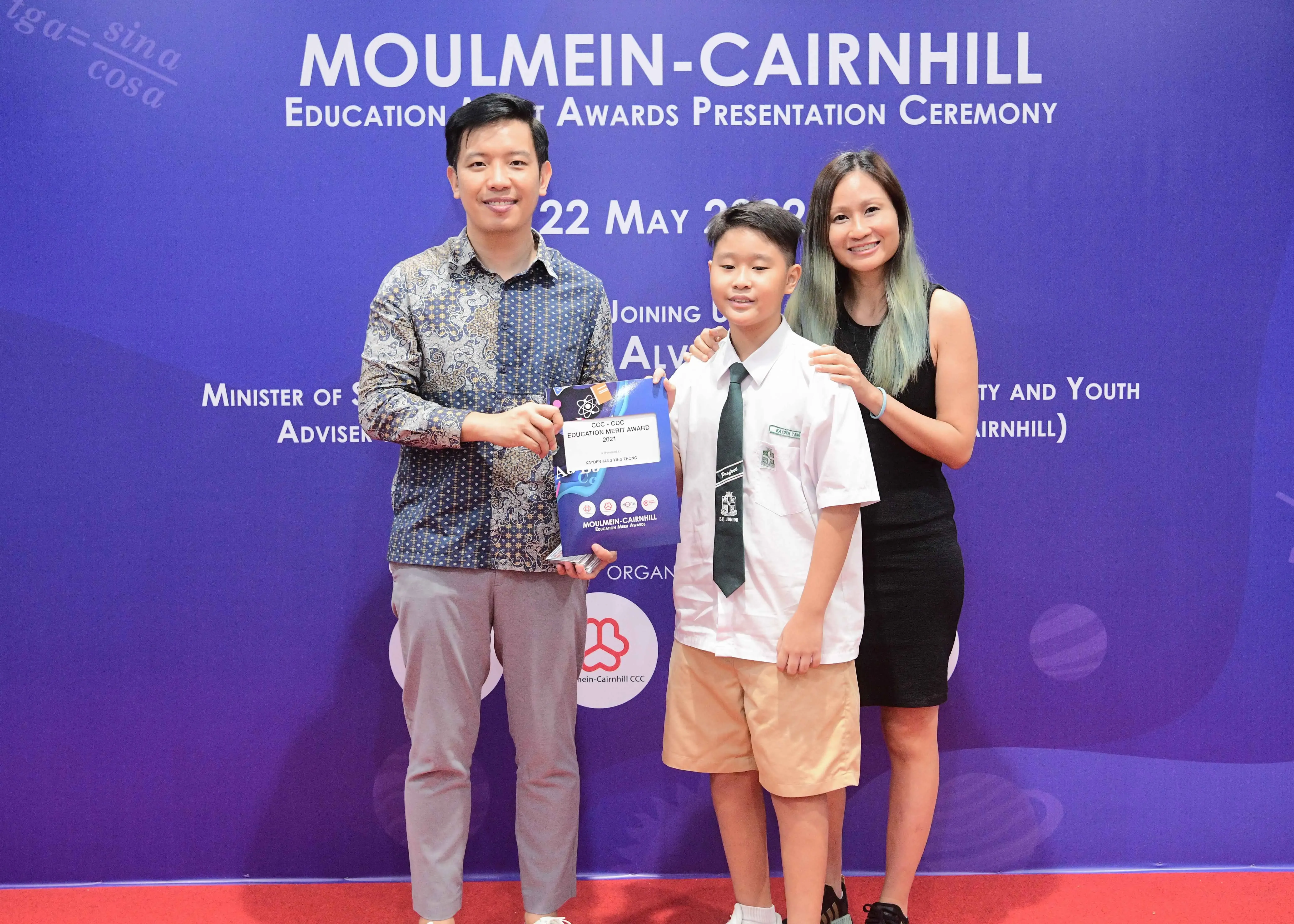 A man, a uniformed boy holding an award, and a woman pose at the Moulmein-Cairnhill Education Merit Awards Ceremony.