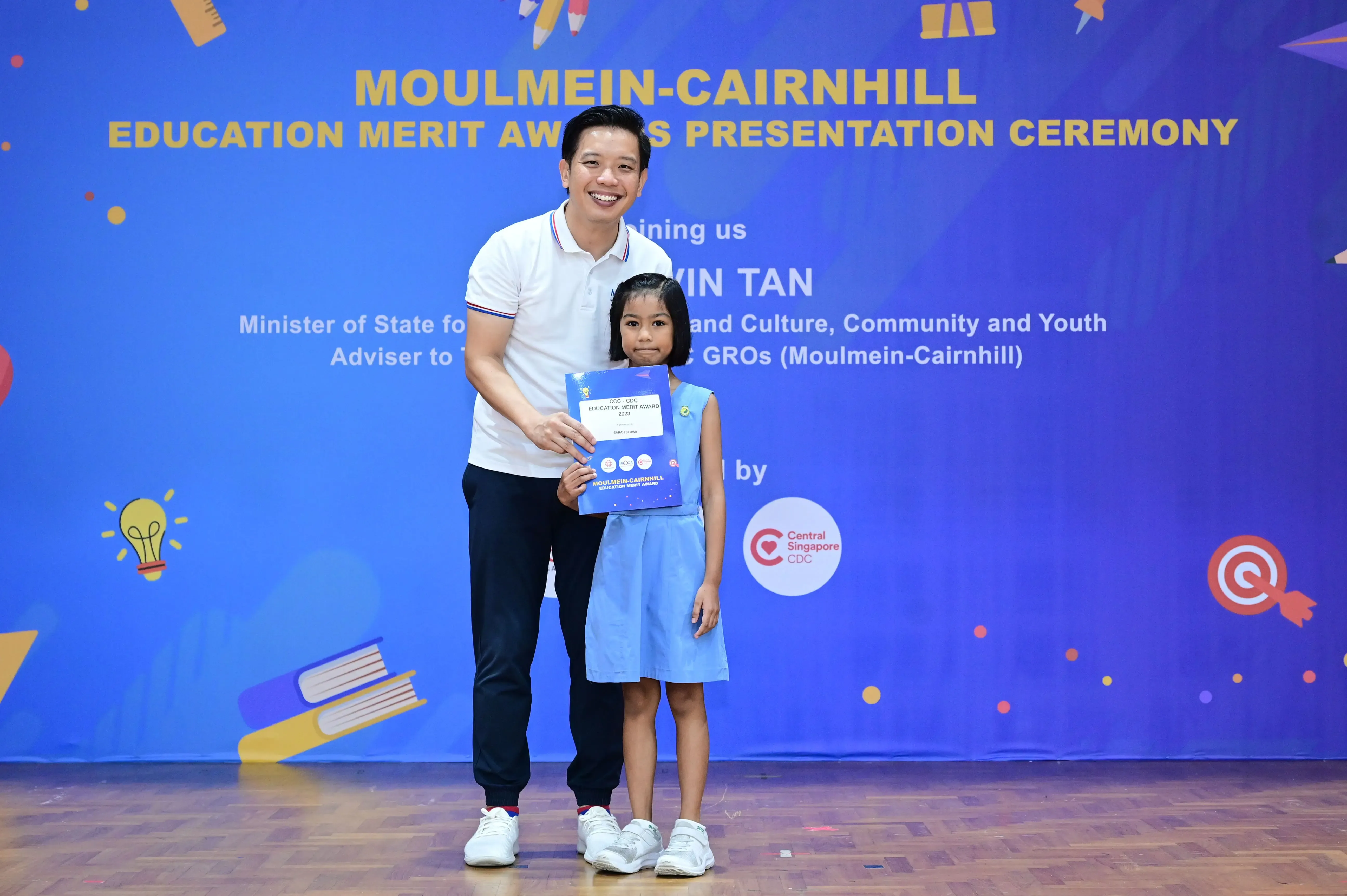 A man and a young girl hold a certificate on stage at the Moulmein-Cairnhill Education Merit Awards ceremony.