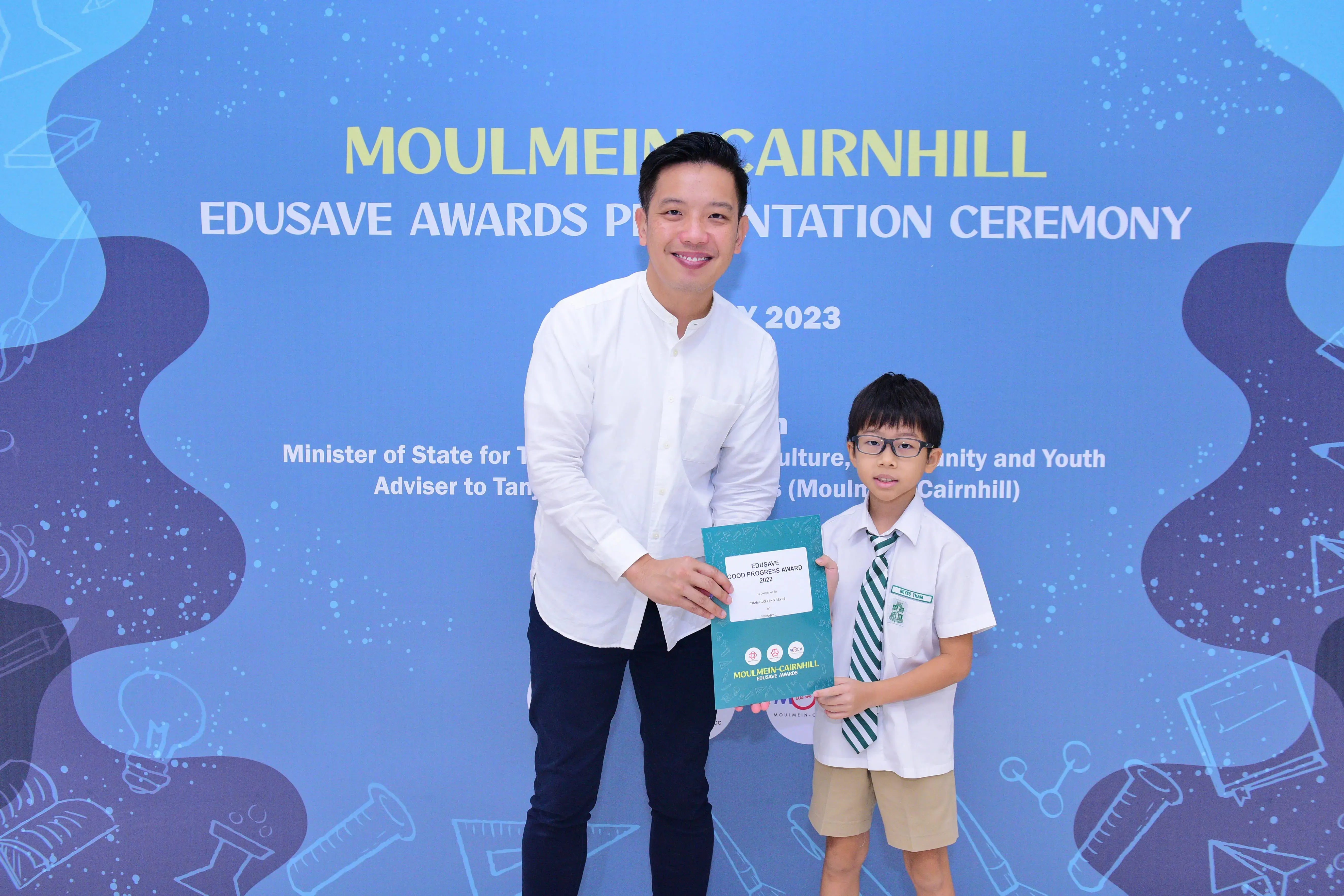 A man and a young boy in school uniform hold an award certificate at the Moulmein-Cairnhill Edusave Awards Ceremony 2023.