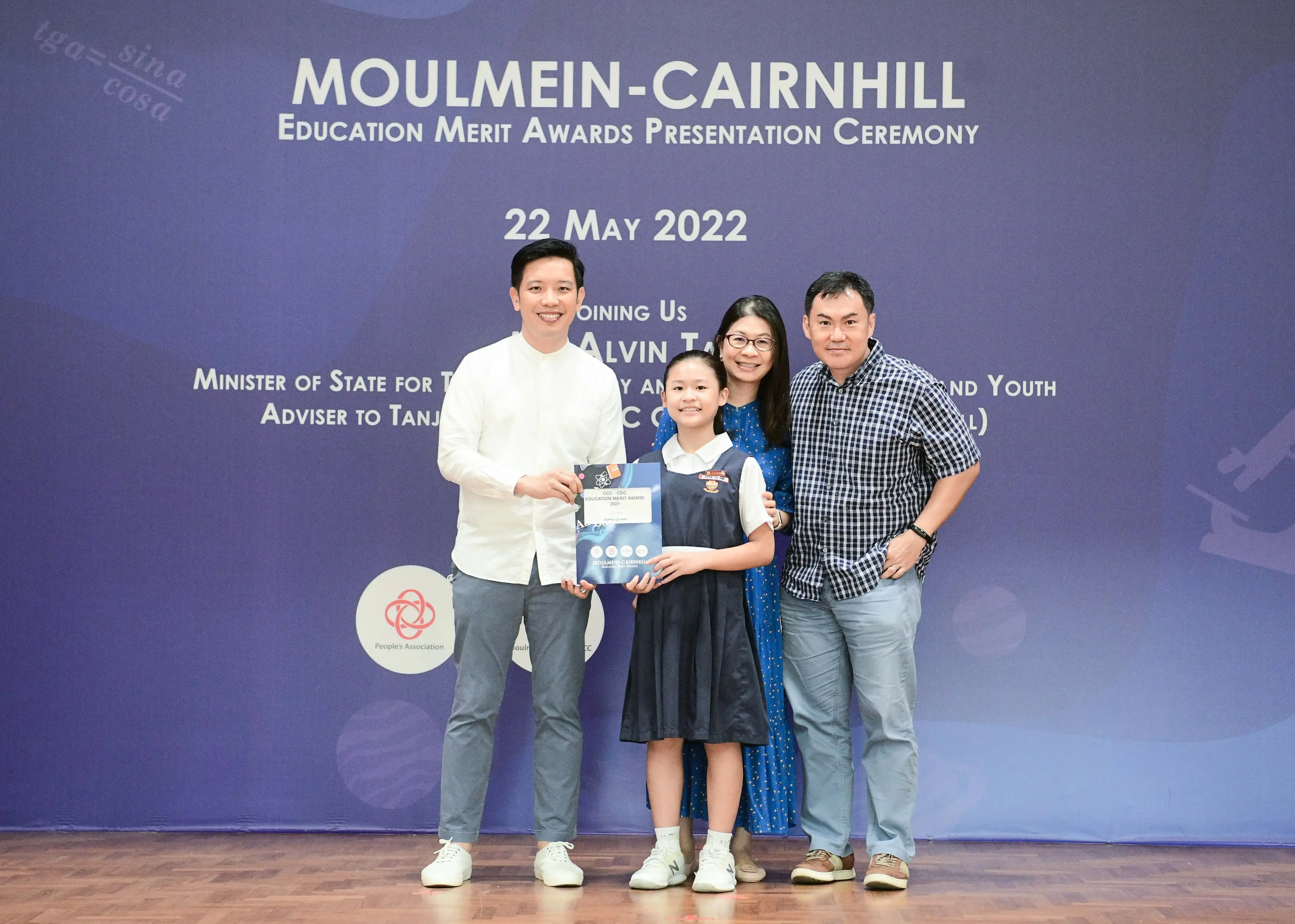 Four people pose in front of a Moulmein-Cairnhill Education Merit Awards banner dated 22 May 2022, with a girl holding an award.