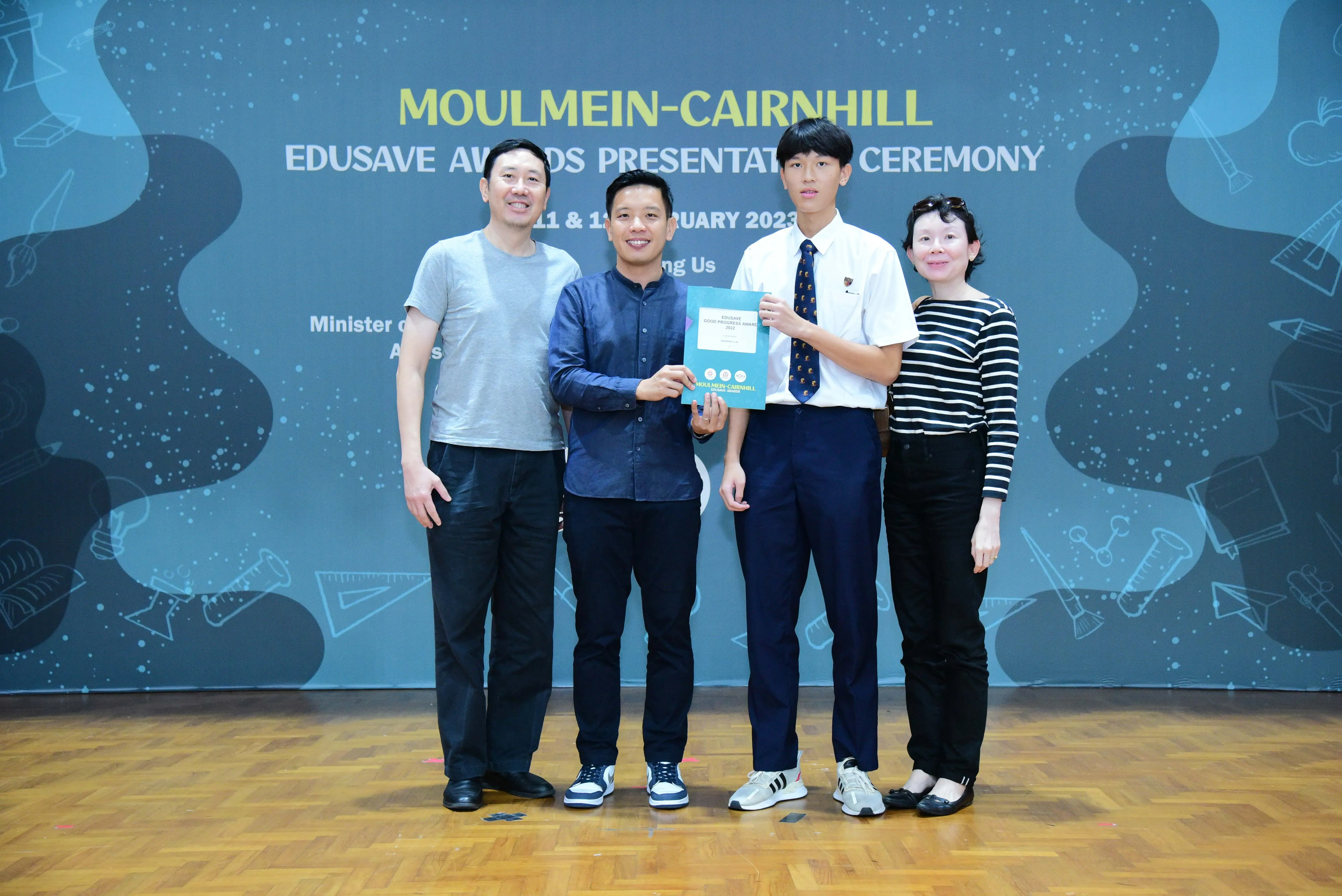 Four people stand before a Moulmein-Cairnhill Edusave Awards Presentation Ceremony backdrop, holding a certificate.