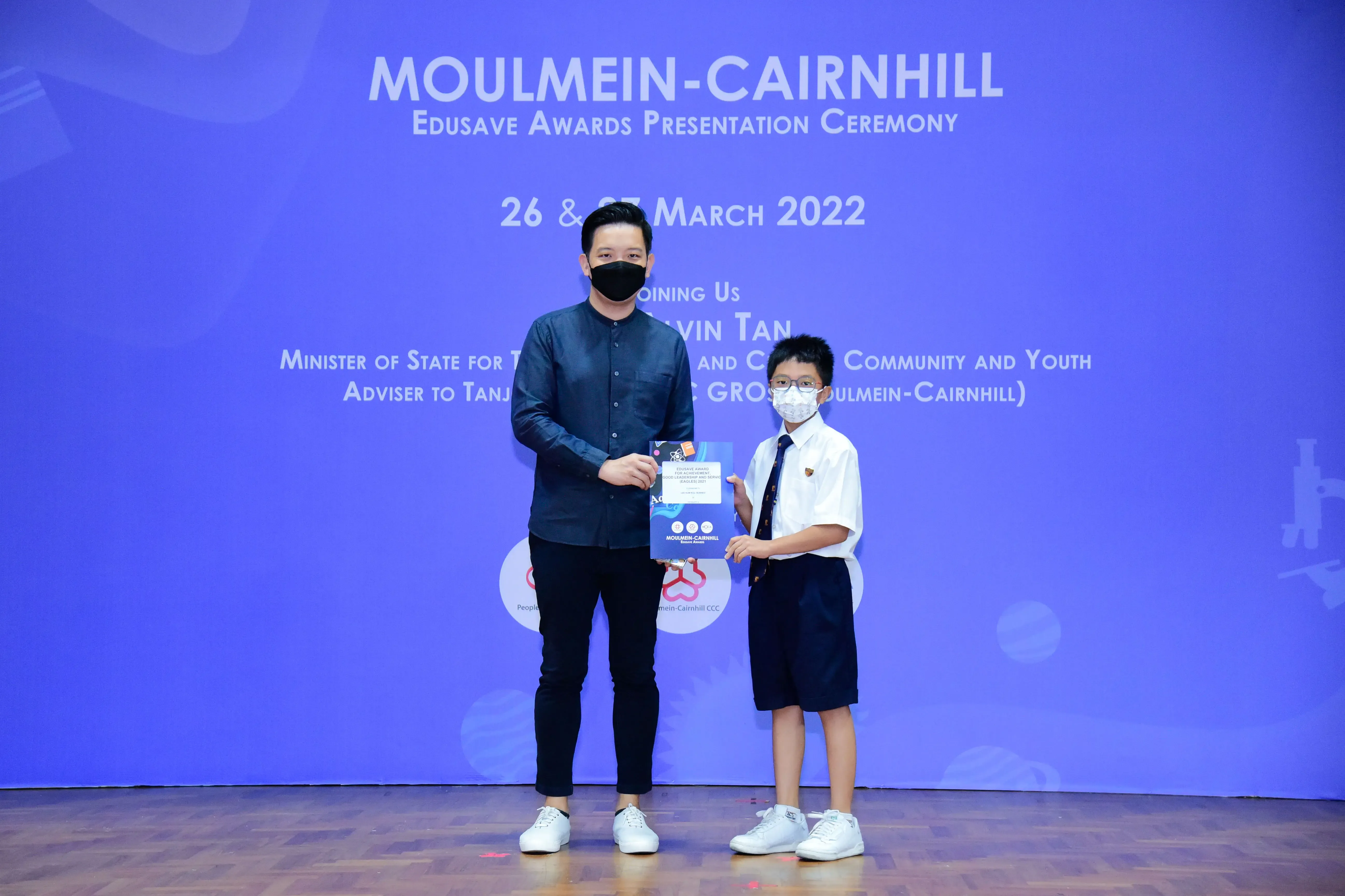 An adult and a young student in school uniform hold an award together at the Moulmein-Cairnhill Edusave Awards Ceremony 2022.