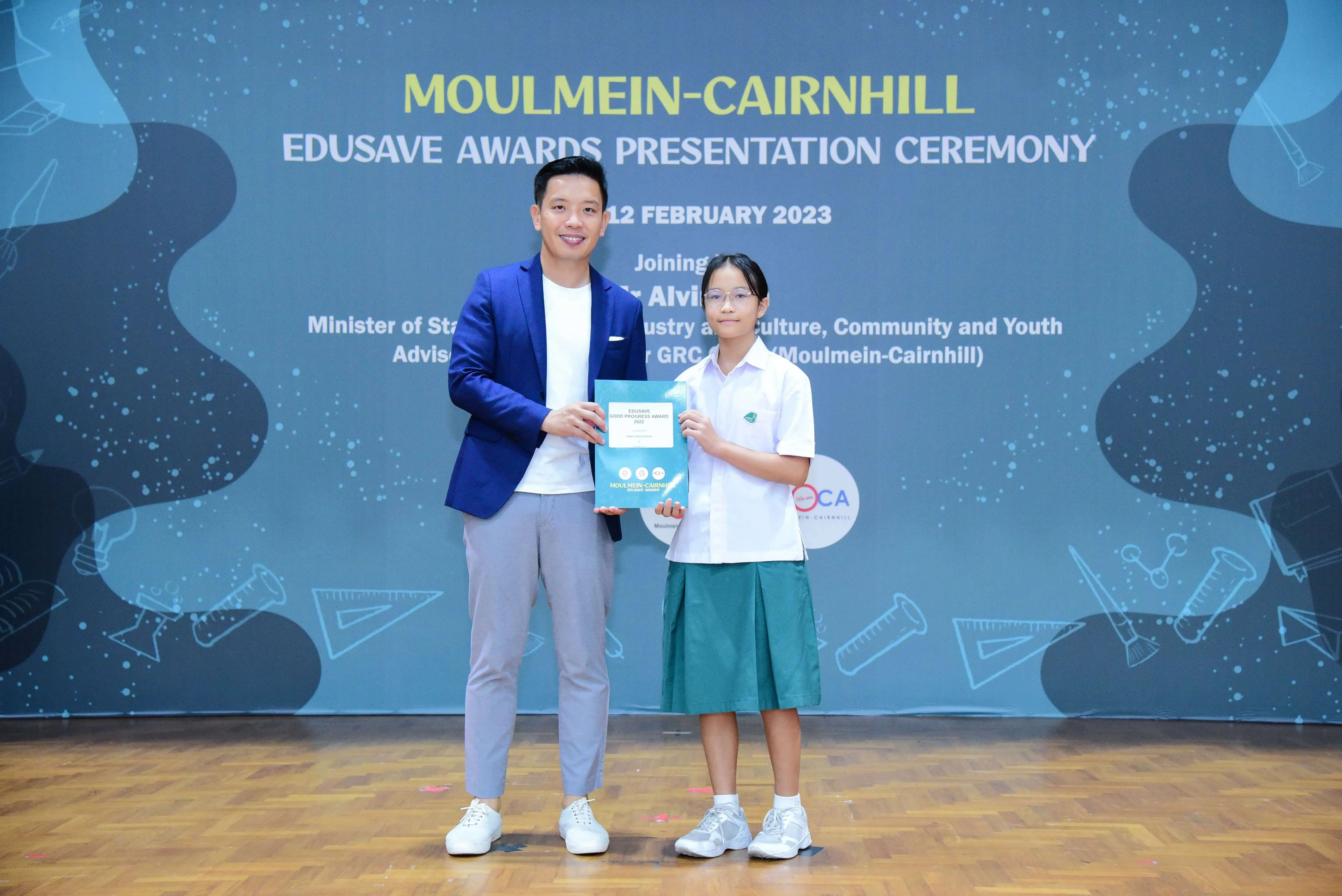 A man in a blue blazer and a young girl in school uniform hold a certificate at the Moulmein-Cairnhill Edusave Awards 2023.