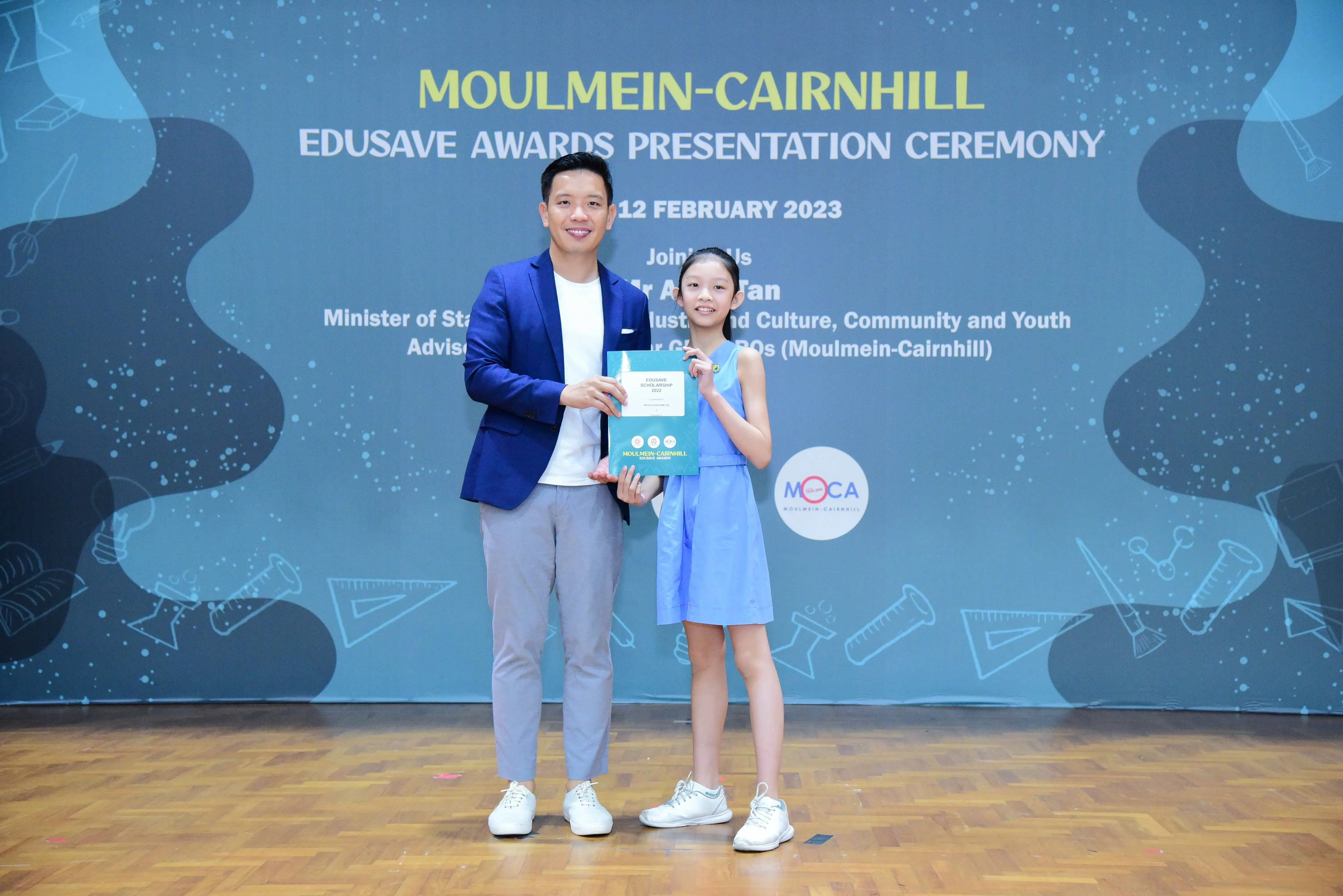 A man in a blue blazer and a young girl in a blue dress hold a certificate at the Moulmein-Cairnhill Edusave Awards Ceremony.