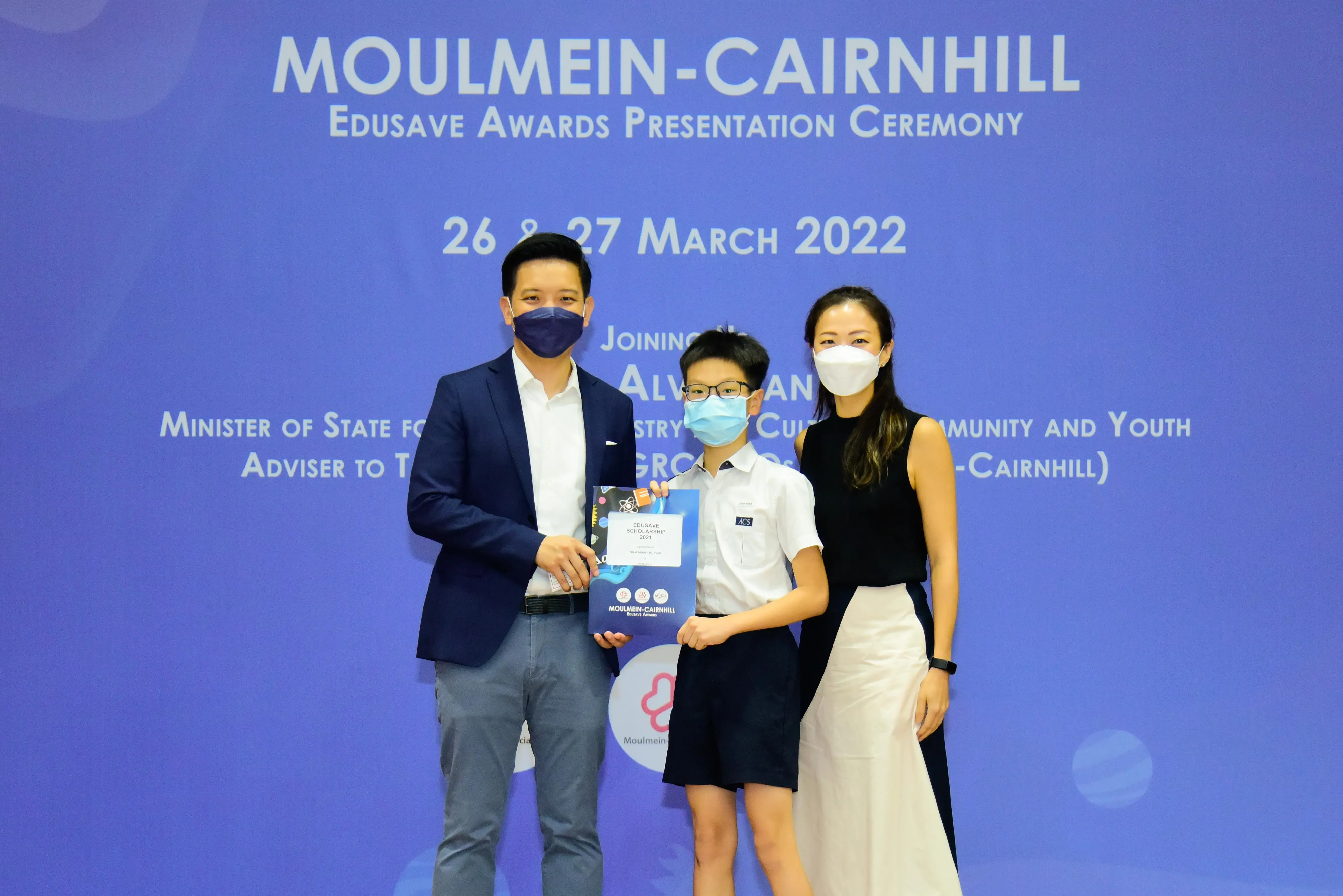 Three masked people pose at the Moulmein-Cairnhill Edusave Awards Ceremony on 26-27 March 2022, holding an award.